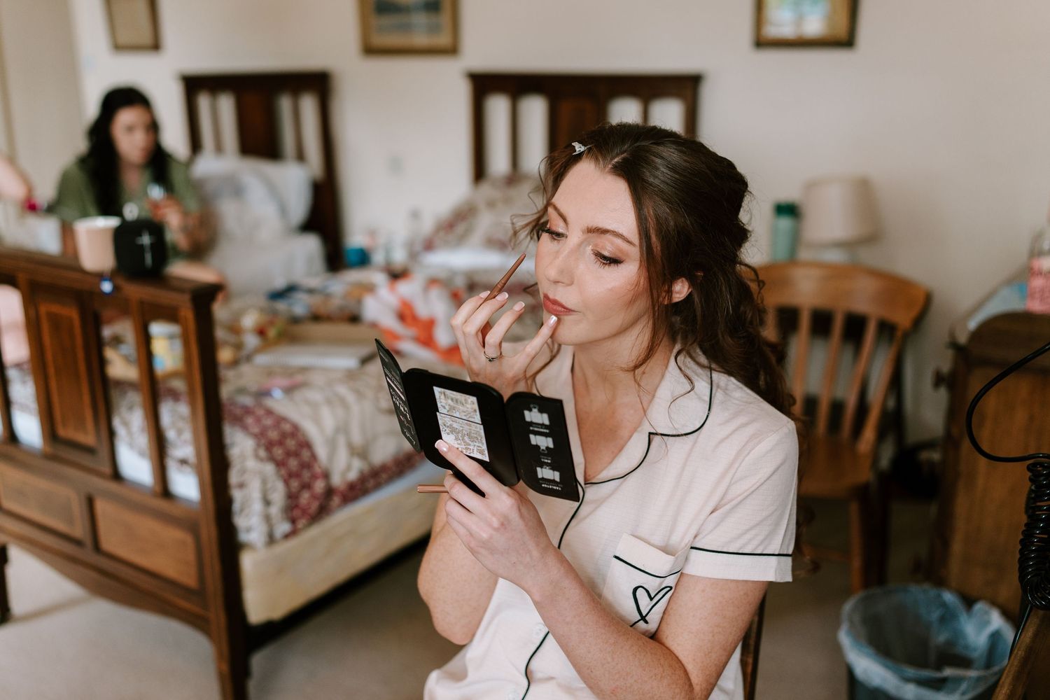 Bride in white pajamas sipping champagne while reading a book during wedding morning preparations.