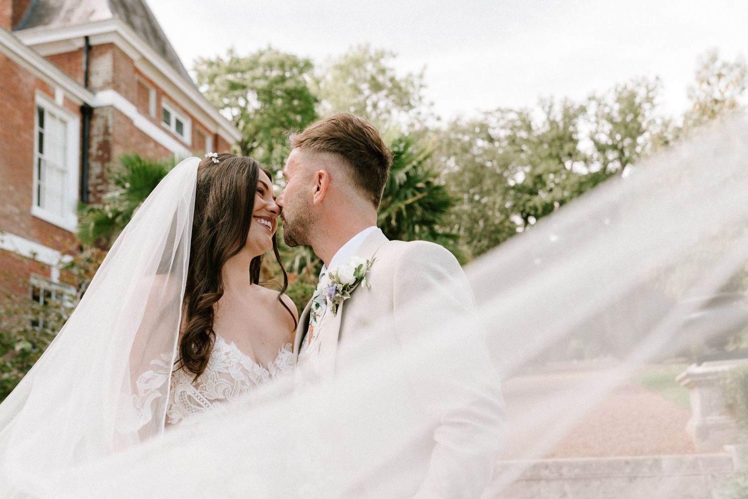 Bride and groom share a romantic kiss outdoors, with the wedding veil flowing dramatically in the wind.