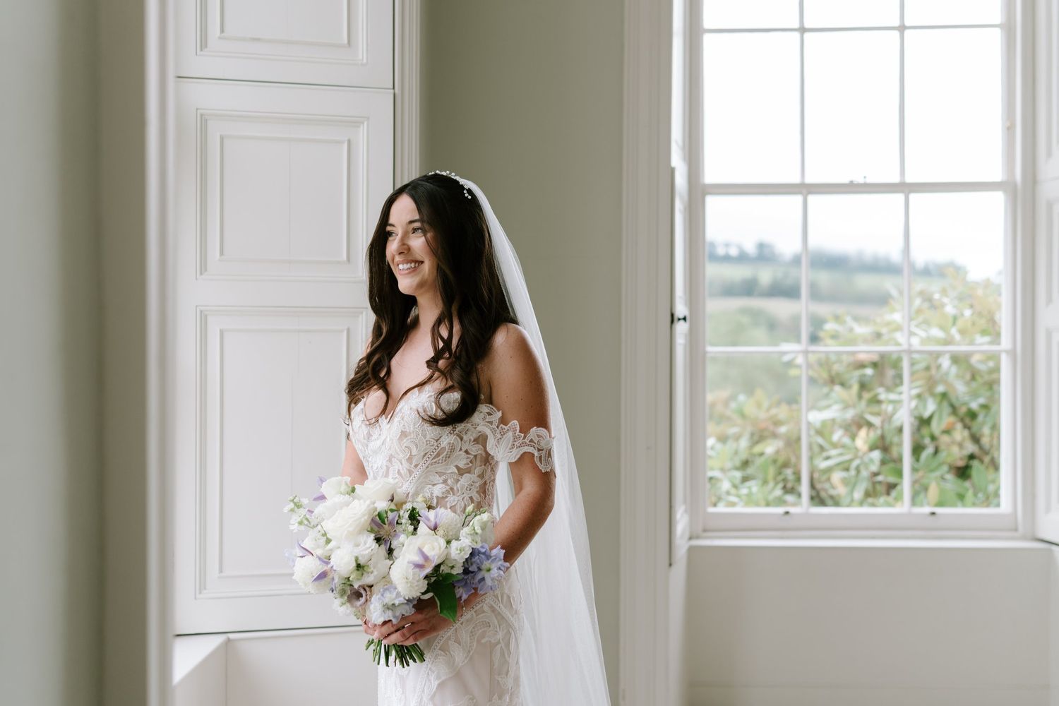 Bride in lace wedding dress and veil holds white floral bouquet, smiling by a bright window in elegant room.