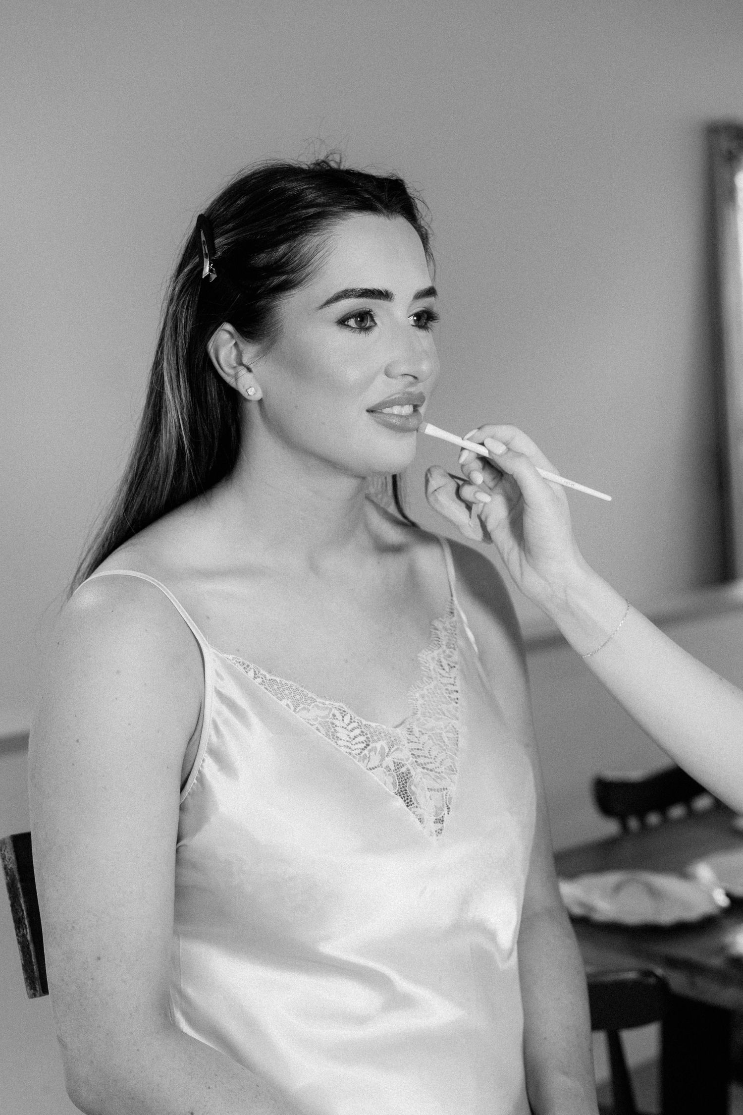 Bride getting lip makeup applied by makeup artist, wearing white satin slip dress, black and white photo.