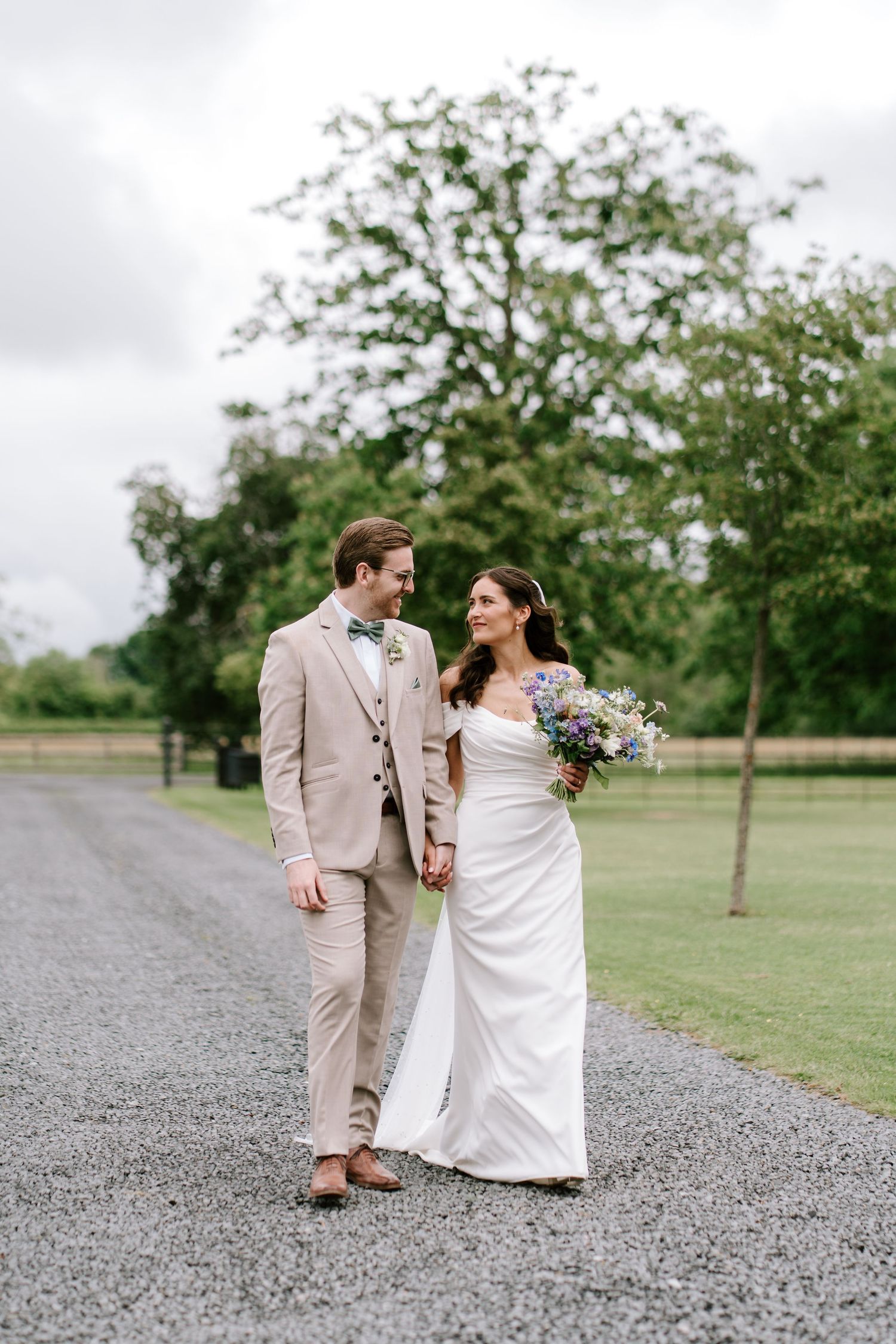 Bride holding wildflower bouquet and groom in beige suit walk hand in hand along tree-lined gravel path.