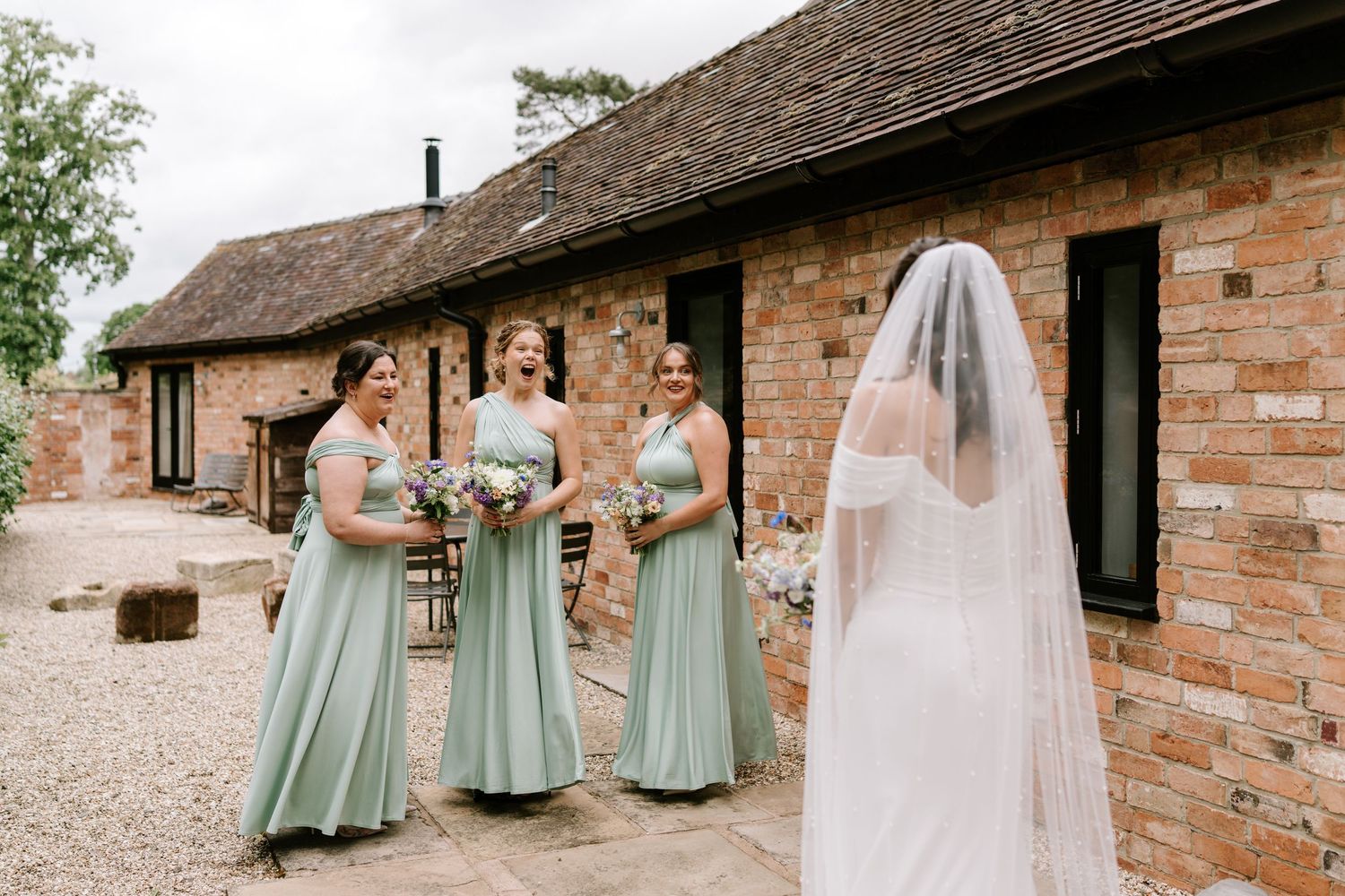 Three bridesmaids in sage green dresses reacting to bride in white veil outside rustic brick barn.