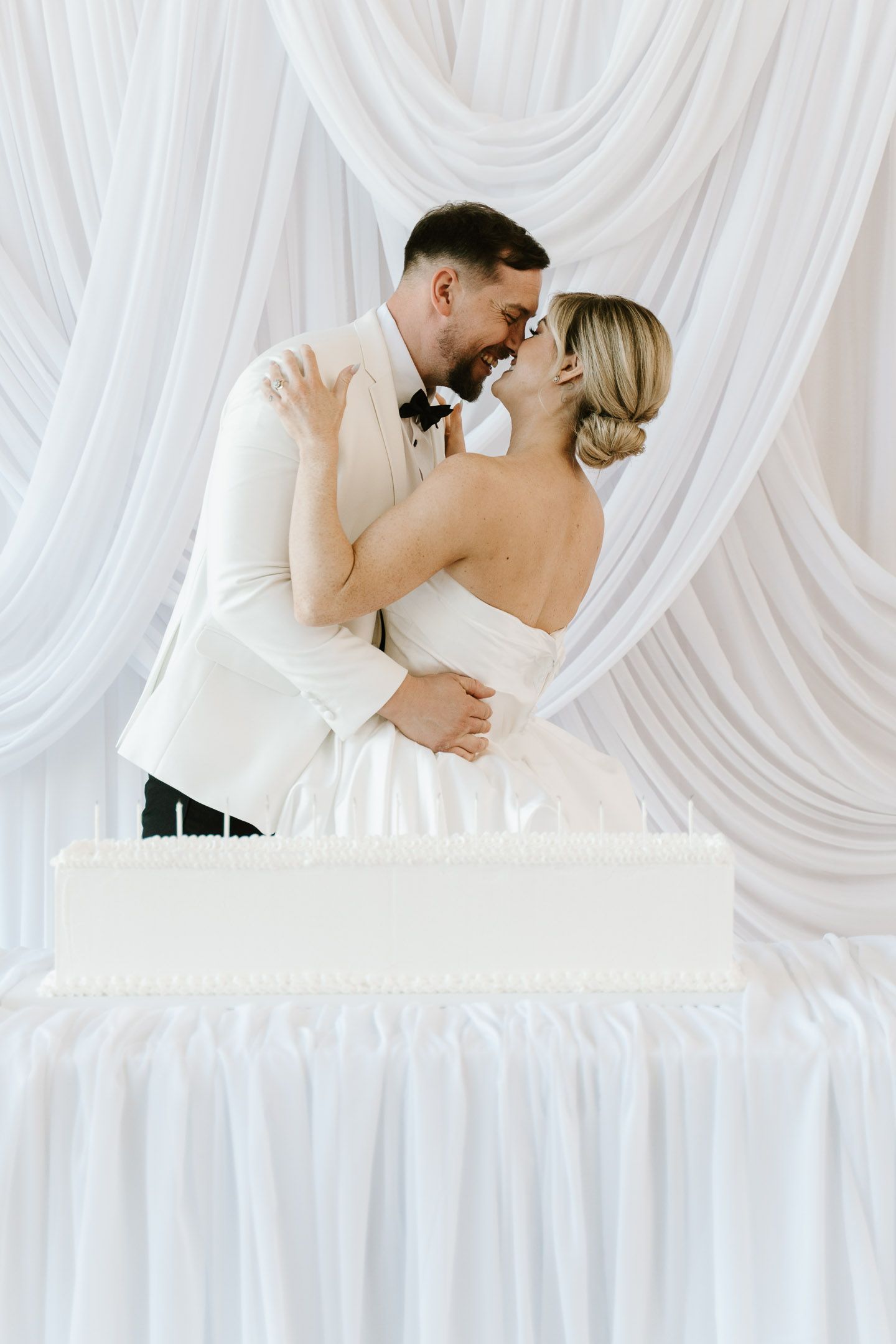 Bride and groom sharing a romantic moment behind a white wedding cake with elegant draped fabric backdrop.