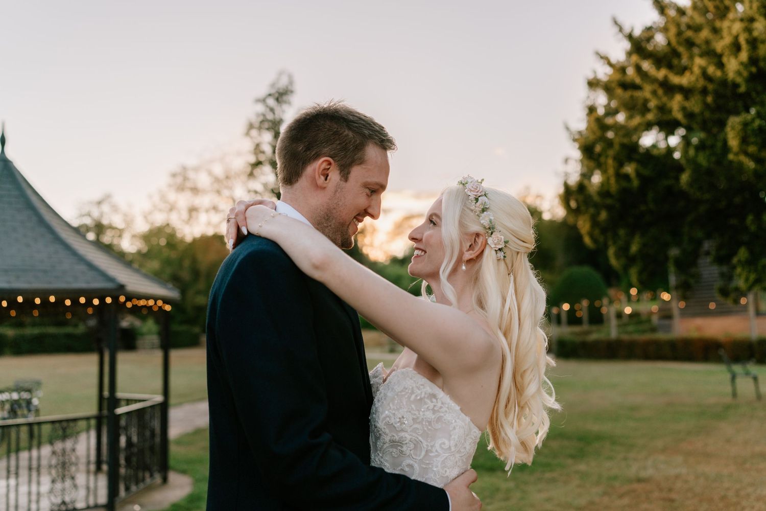 A bride and groom share a romantic moment at sunset in a lush garden, with the bride in a lace gown and floral hair.