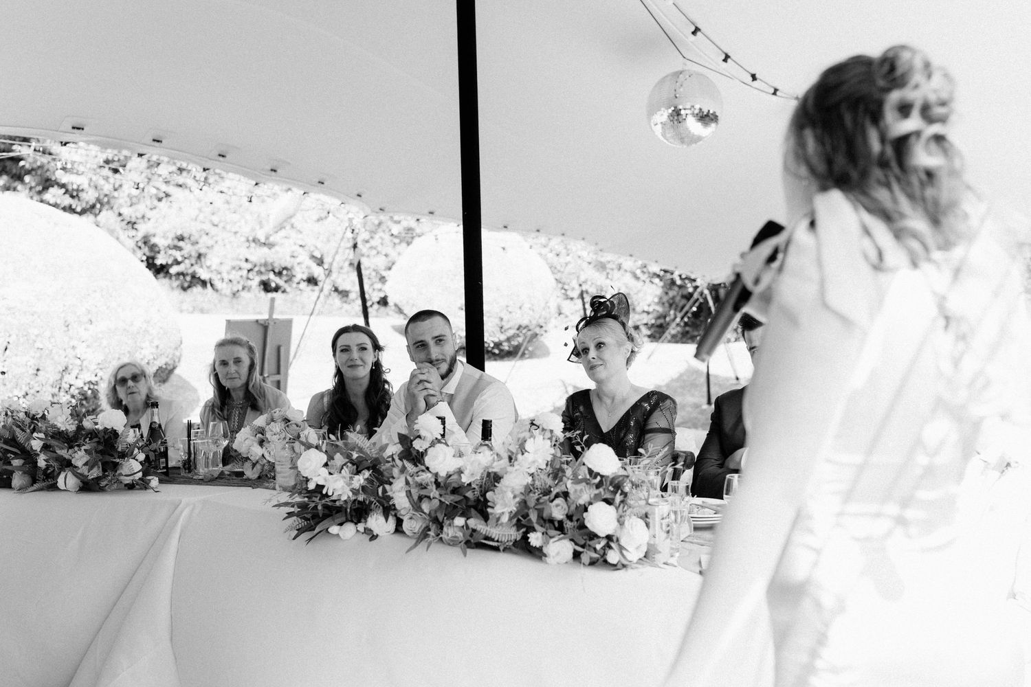 Black and white wedding reception photo showing bridal party at head table with floral arrangements while guest speaks.