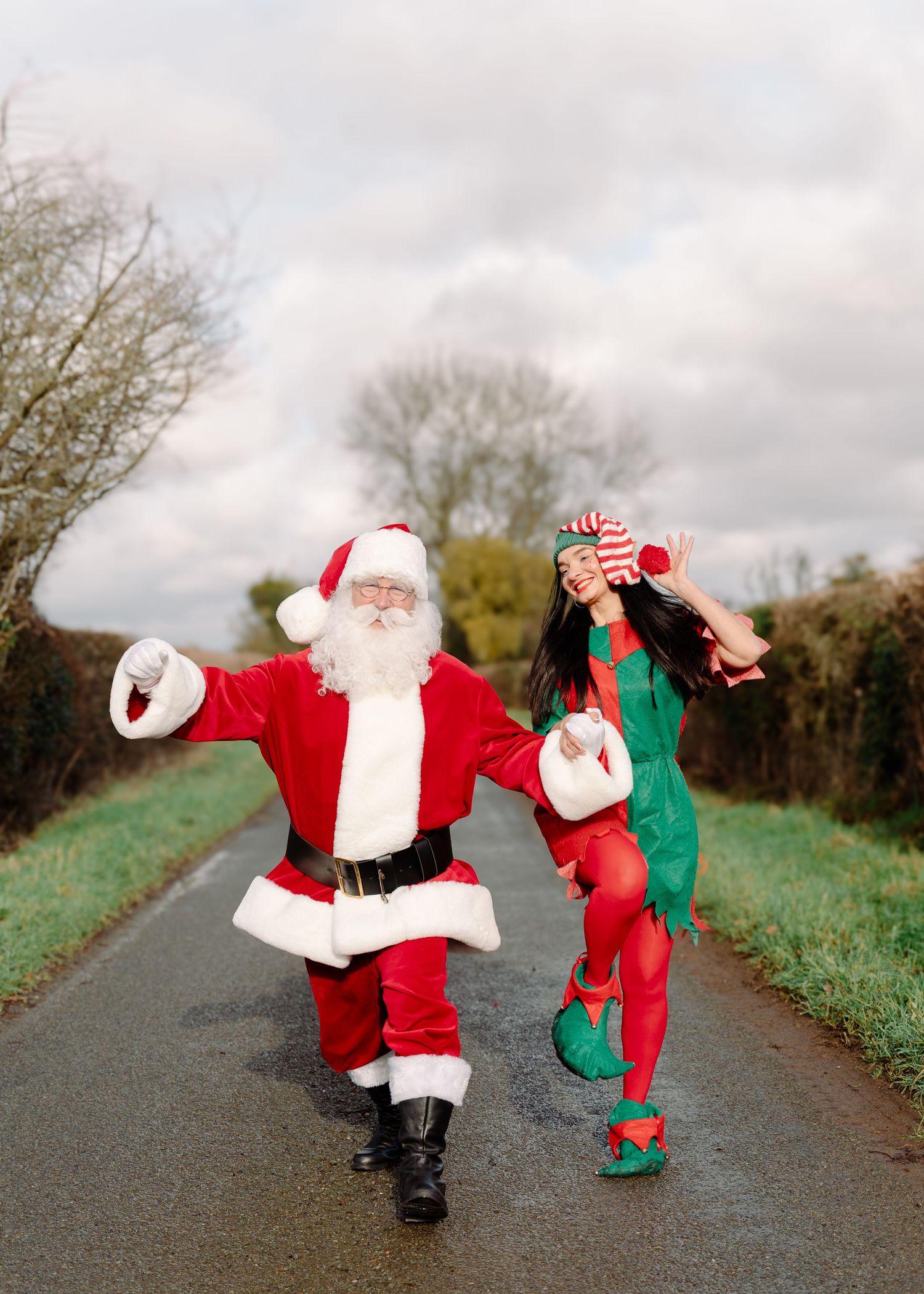 Santa Claus and an elf dance joyfully on a rural road surrounded by winter trees.