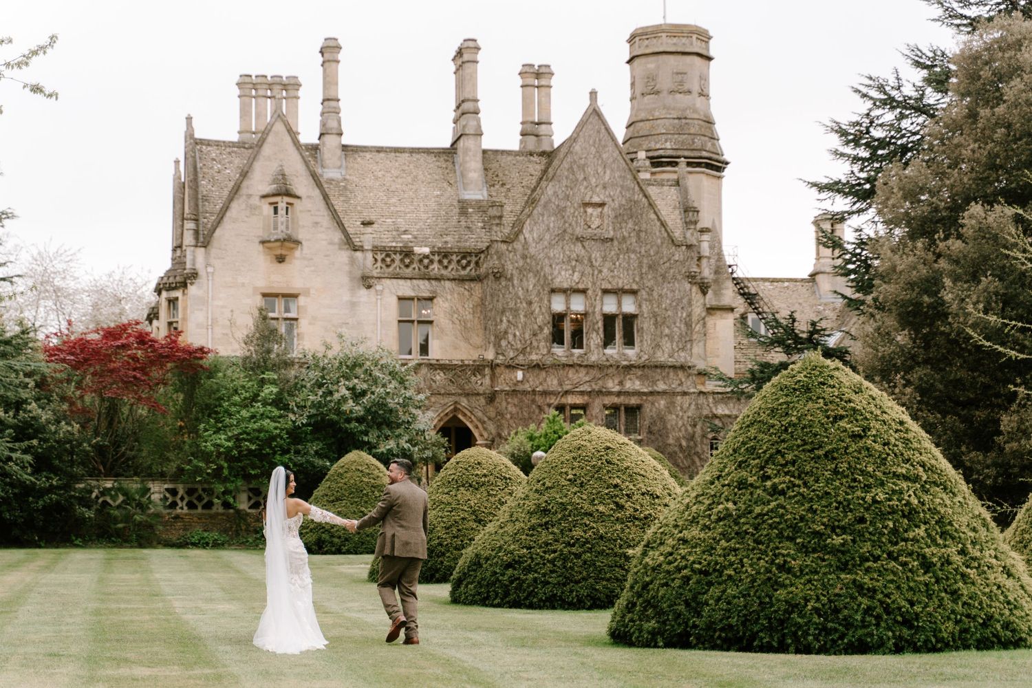 Bride and groom dancing on manicured lawn before grand English manor house with topiary hedges.