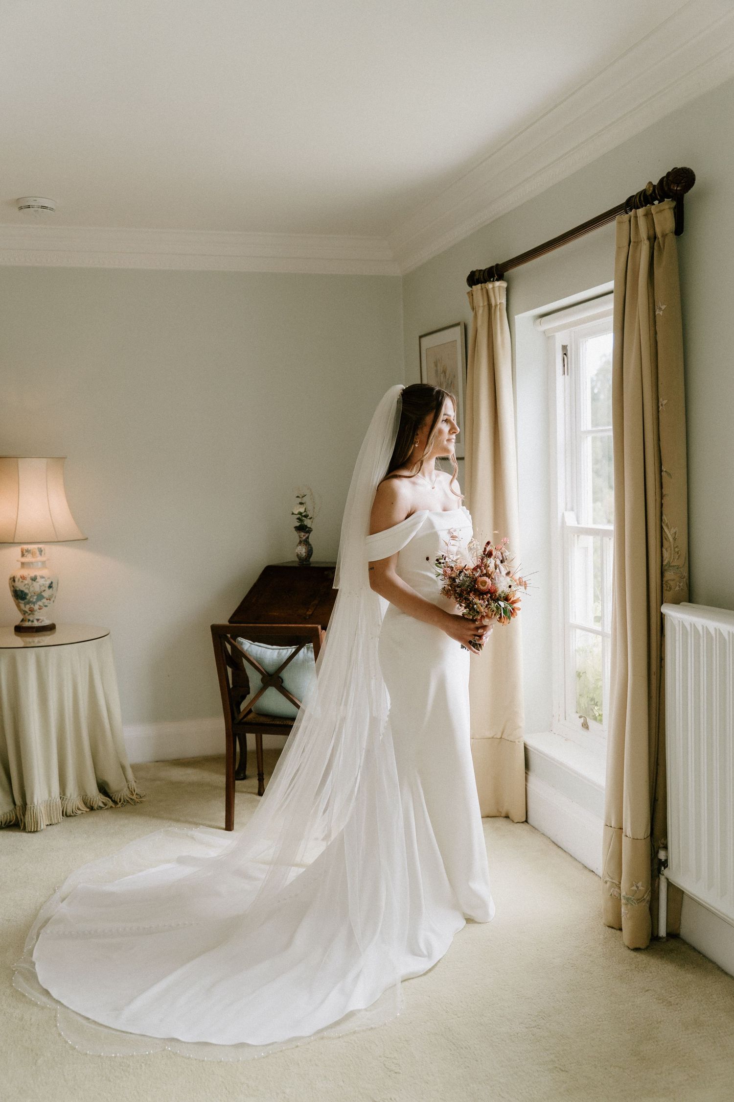 Birtsmorton Court Bride in white off-shoulder gown with long veil holds bouquet by window in elegant room.