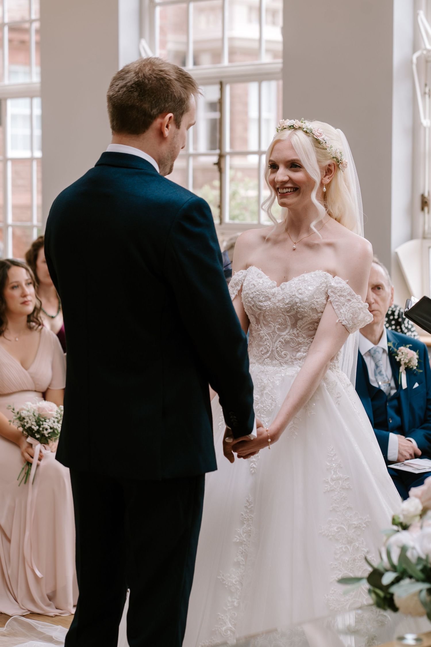 Bride in off-shoulder lace gown holds hands with groom in navy suit during indoor wedding ceremony.