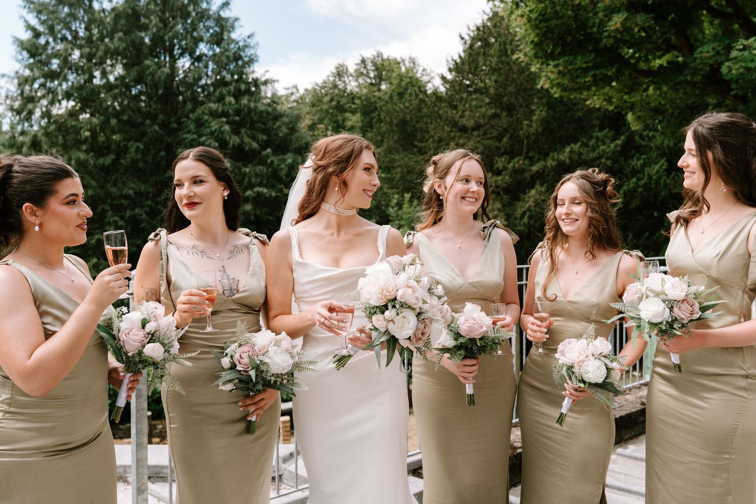 Bride in white gown with bridesmaids in sage green dresses holding bouquets and champagne glasses outdoors.