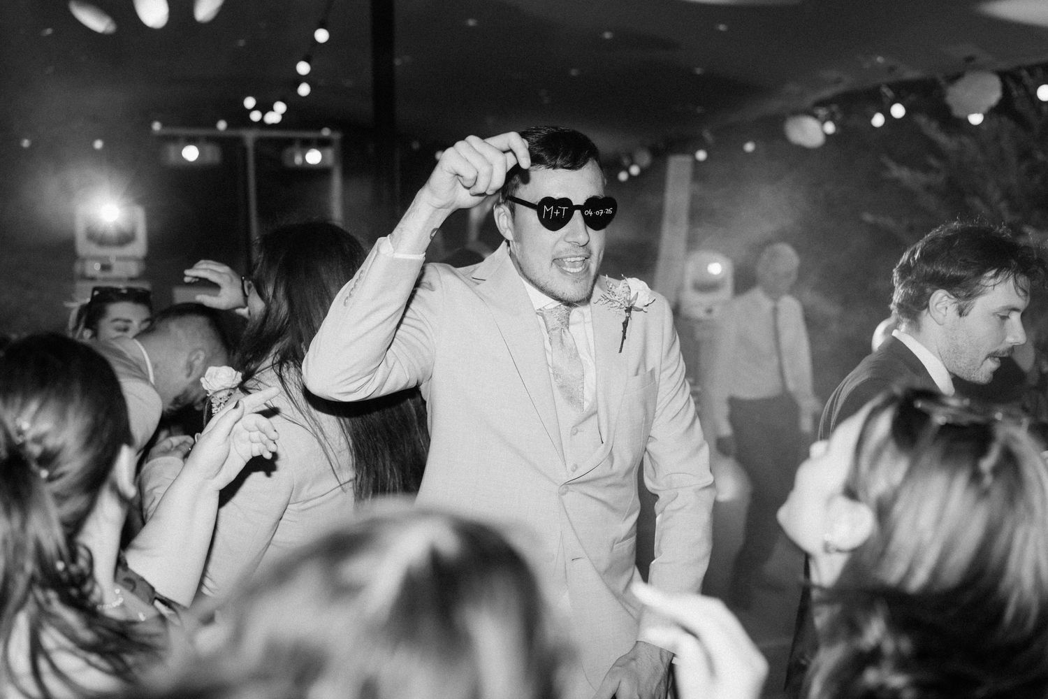 Groom dancing in sunglasses on crowded wedding reception dance floor in black and white.