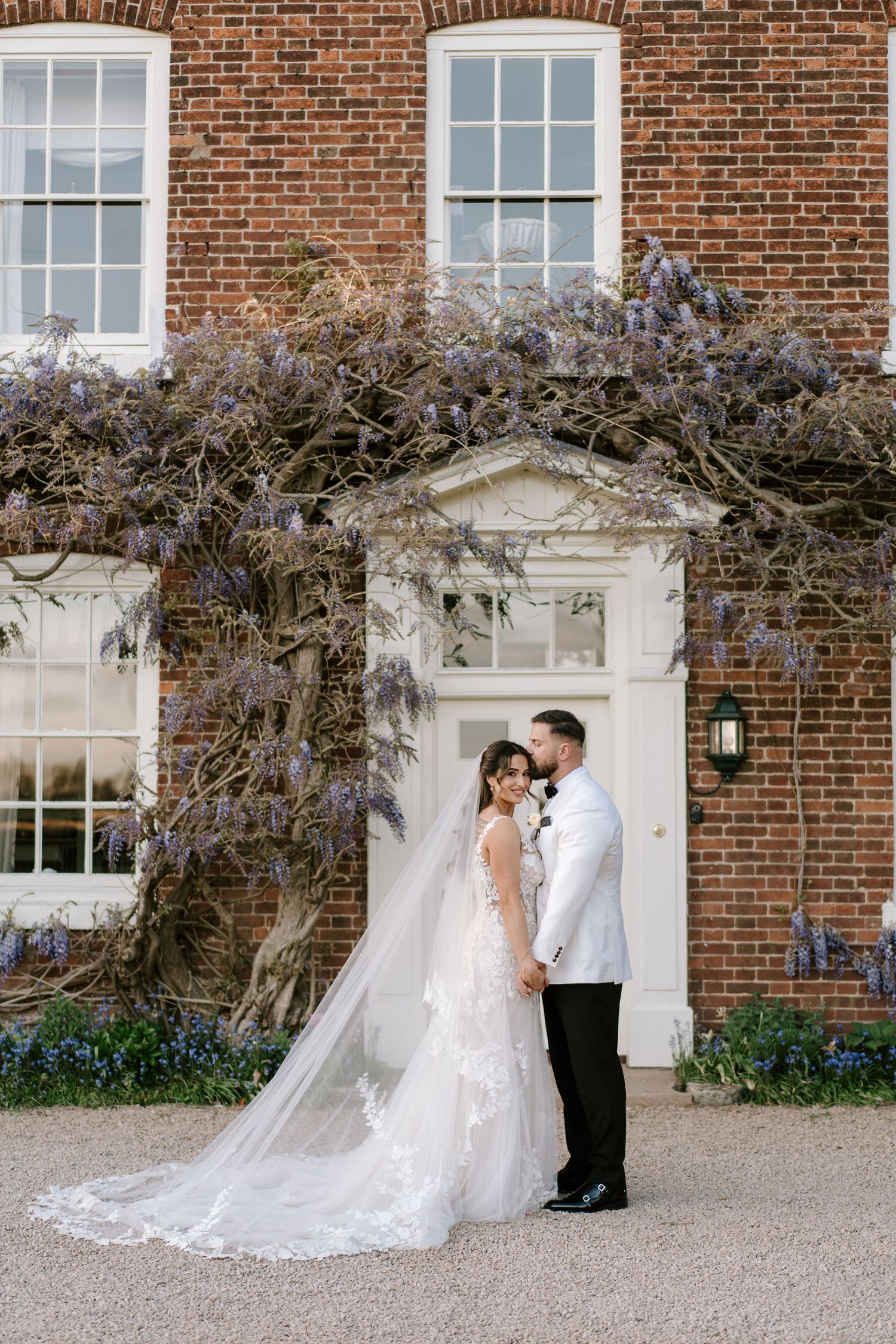 Bride and groom pose before a brick manor adorned with wisteria blooms, her lace gown and cathedral veil trailing elegantly.