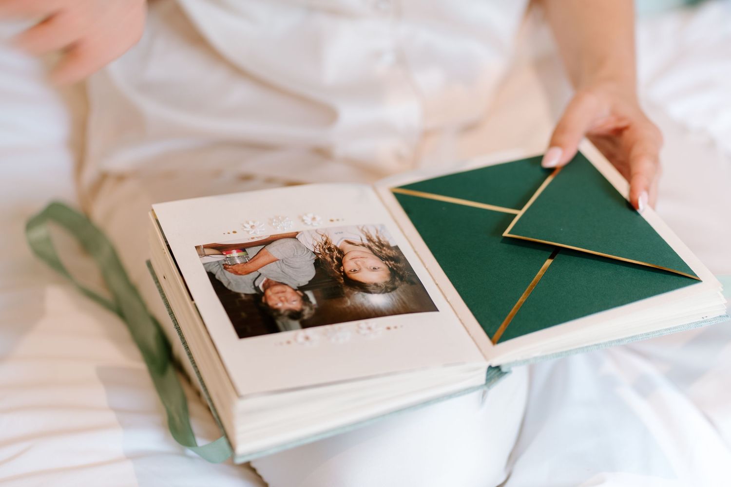 Person holding an open photo album with a family picture and green envelope on a white bed.