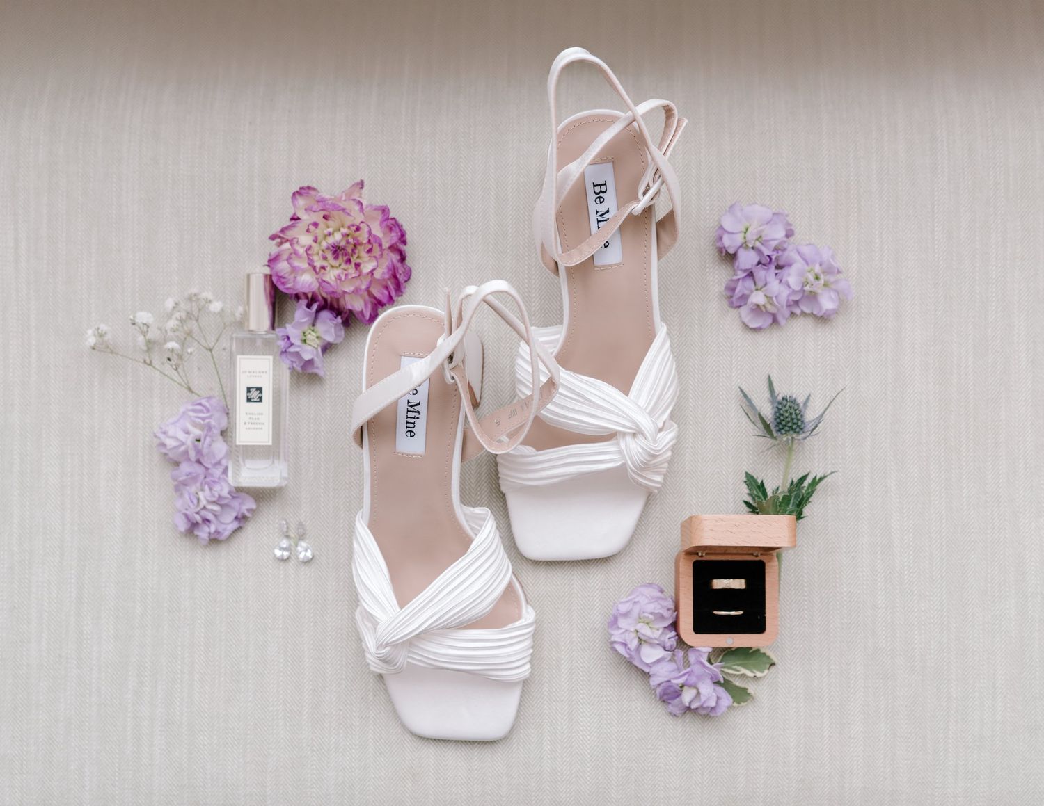 White bridal heels surrounded by purple flowers, a ring box, and perfume on a neutral background.