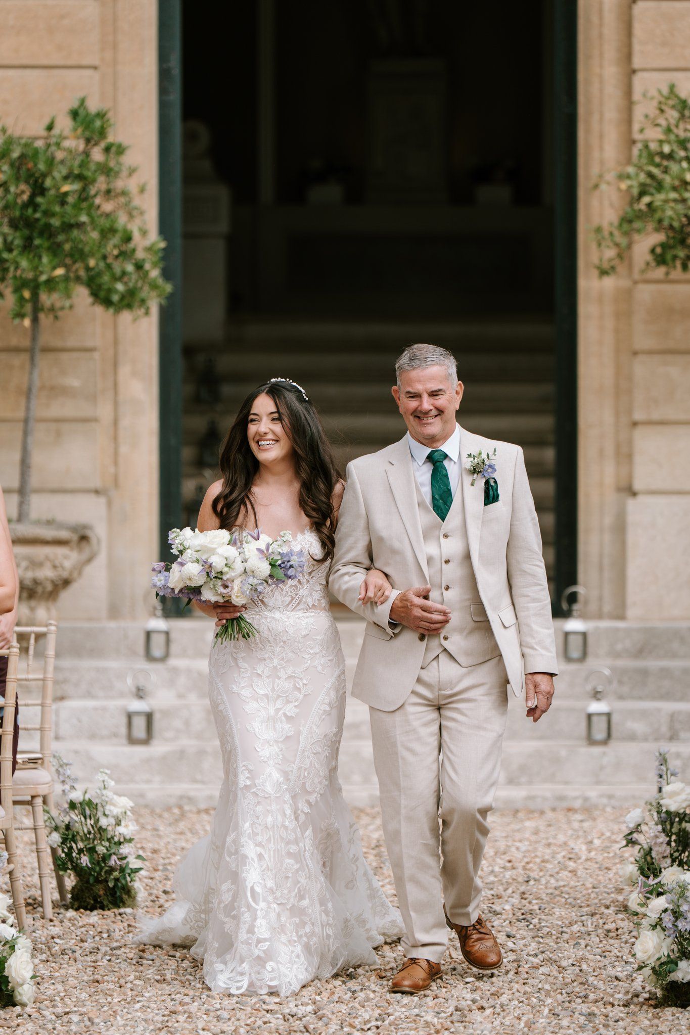 Bride in lace gown walks down aisle with father in beige suit and green tie at outdoor wedding ceremony.
