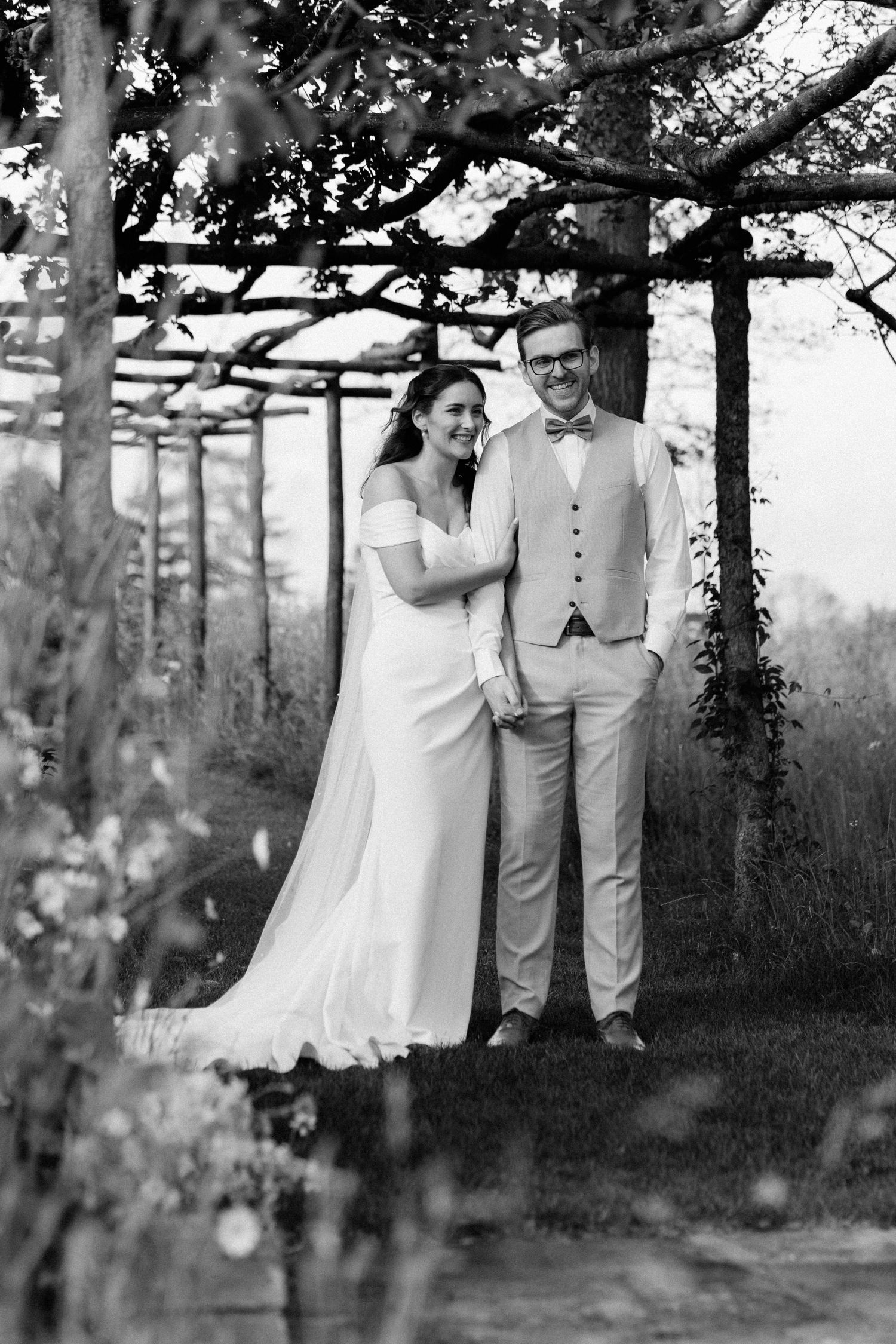 Black and white wedding photo of smiling bride and groom standing under a rustic pergola surrounded by lush greenery.
