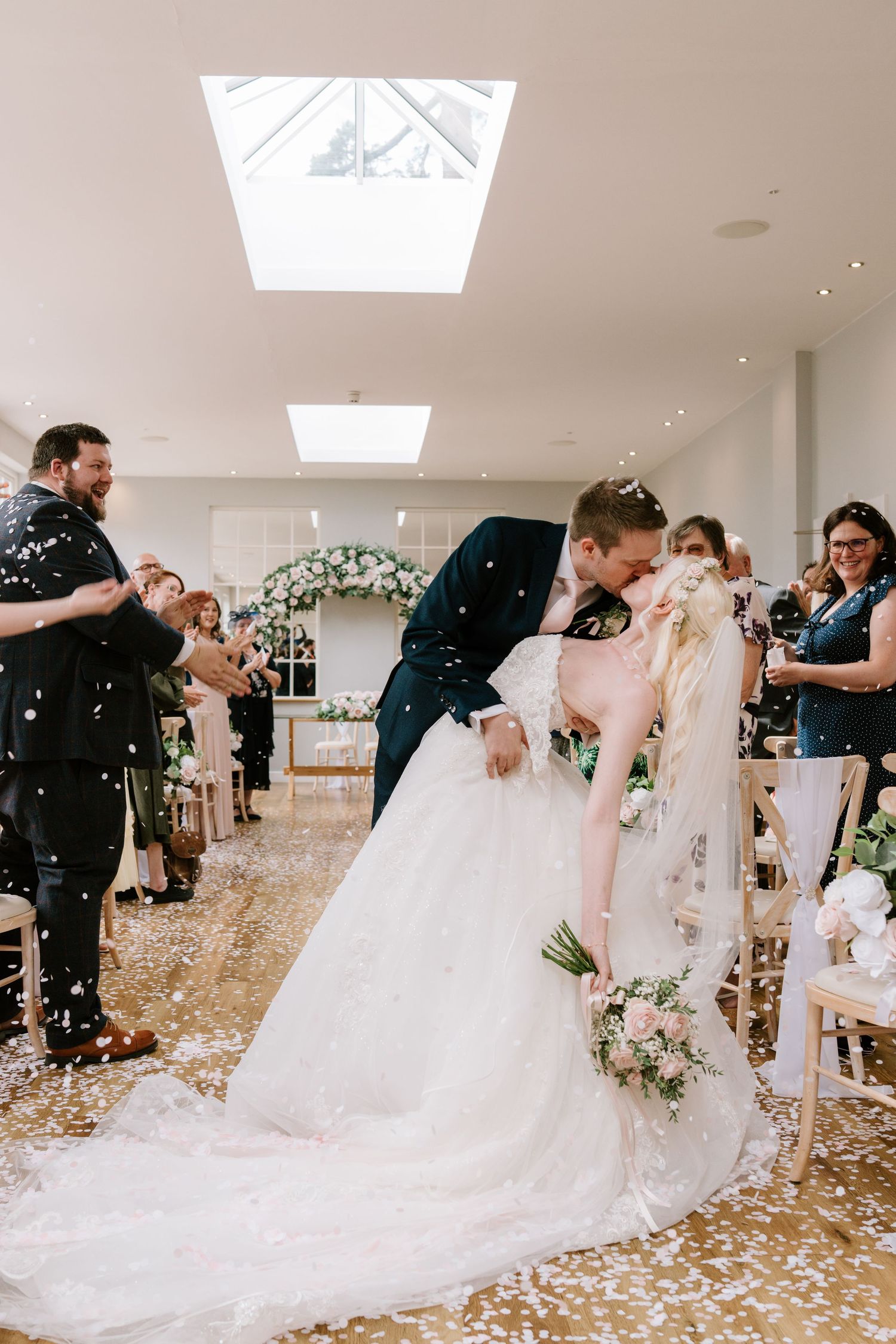 Bride and groom share a romantic dip kiss while confetti falls during their wedding ceremony aisle exit.