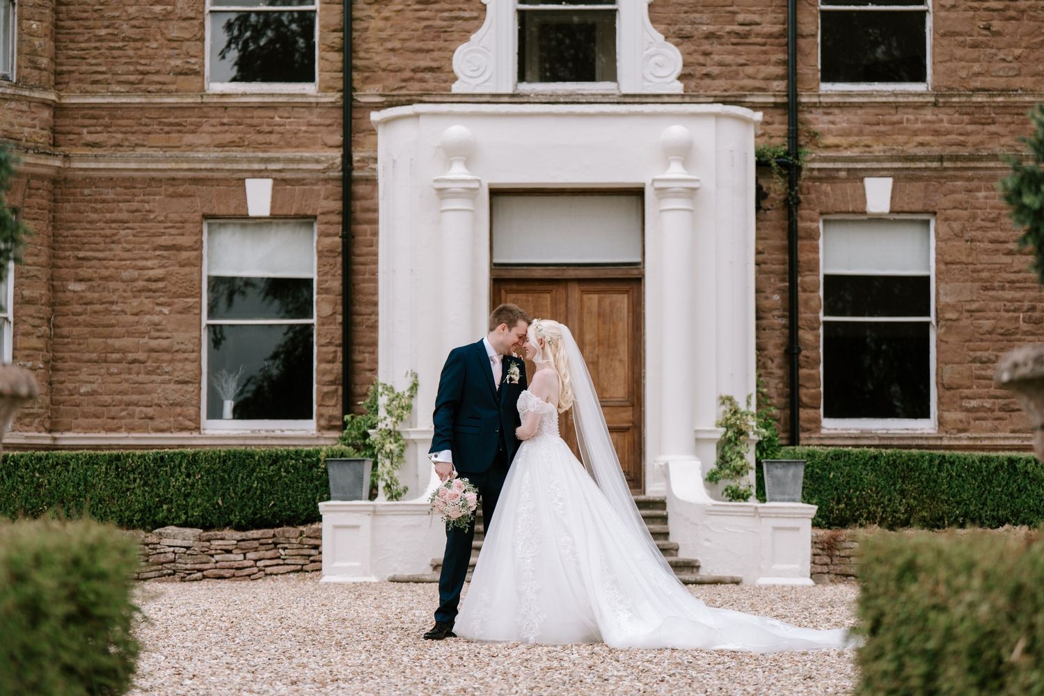 Bride and groom share a romantic kiss outside an elegant brick manor house on their wedding day.