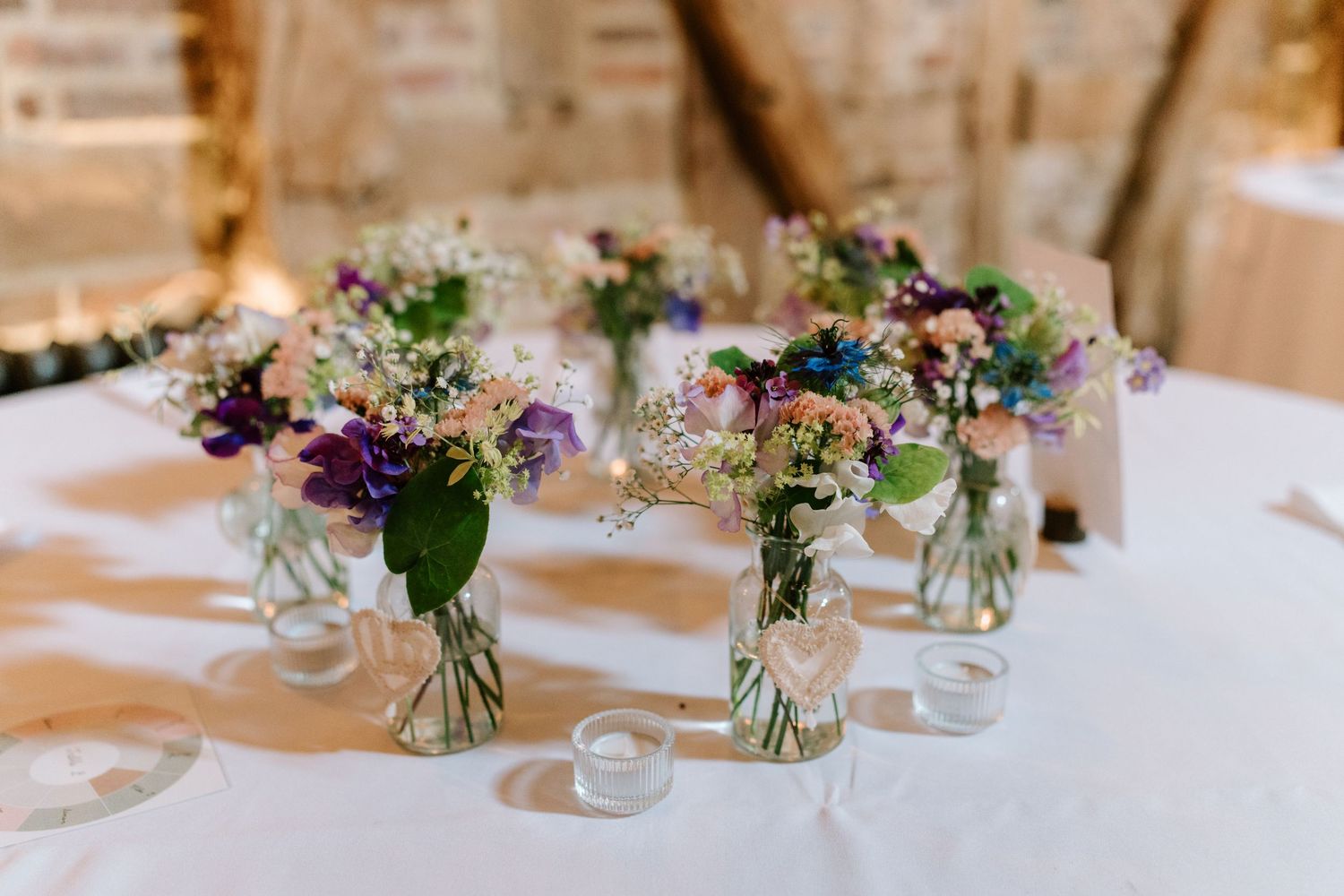 Small glass vases with colorful wildflower bouquets arranged as rustic wedding table centerpieces on a white linen tablecloth.