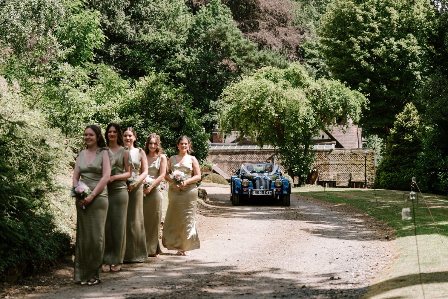 Bridesmaids in sage green gowns walking down sunlit driveway with vintage car and lush trees behind.