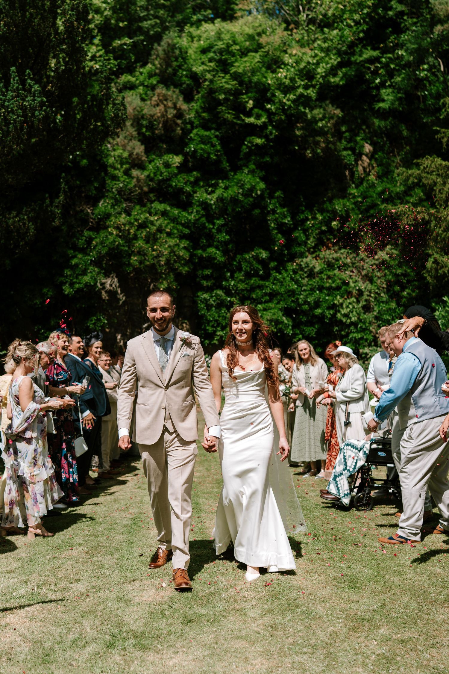 Newlyweds walk hand in hand down aisle as guests celebrate with confetti outdoors.