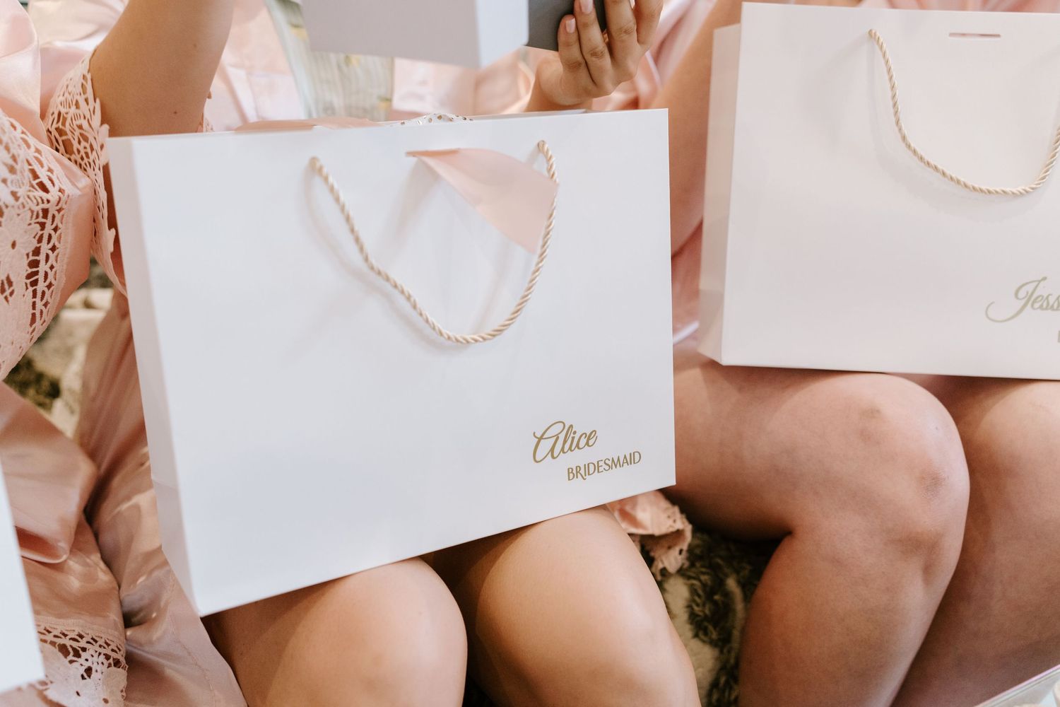 Elegant white gift bags with gold script logo and pink ribbon handles held by women in blush pink robes.