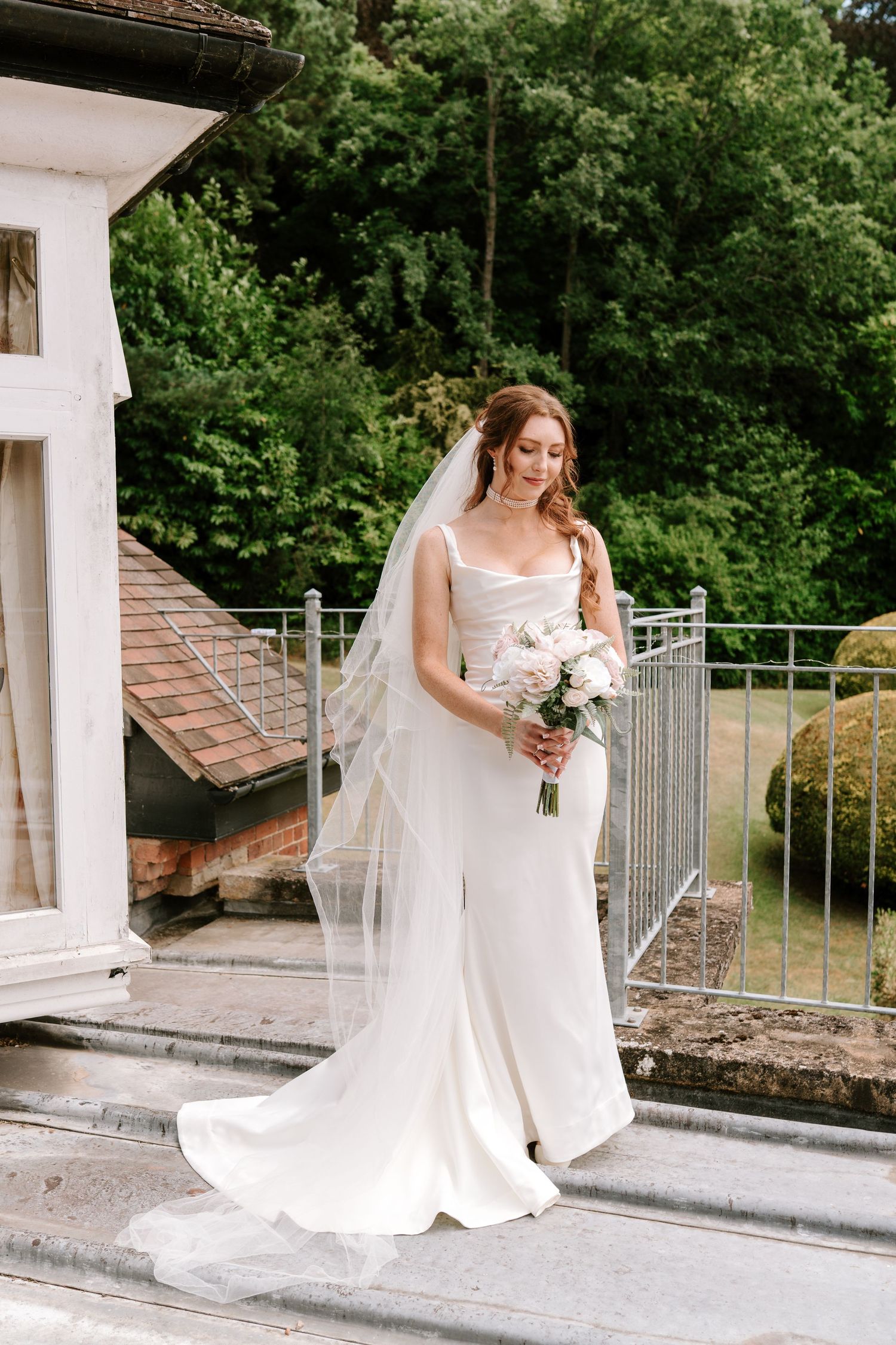 Elegant bride in white gown and veil holding bouquet on outdoor terrace with lush green trees.