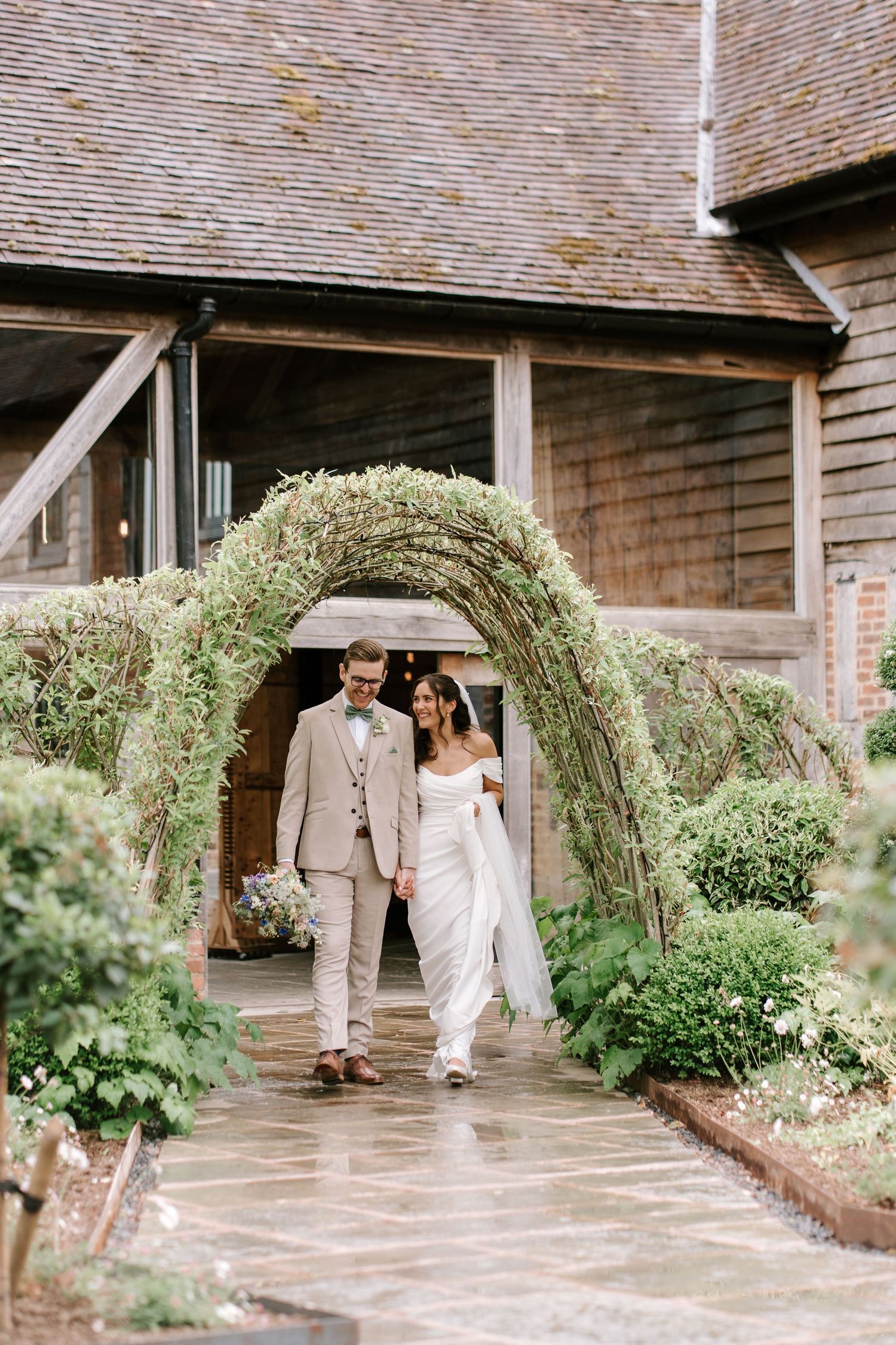 Bride and groom walking hand in hand under lush green archway at elegant rustic barn wedding venue.