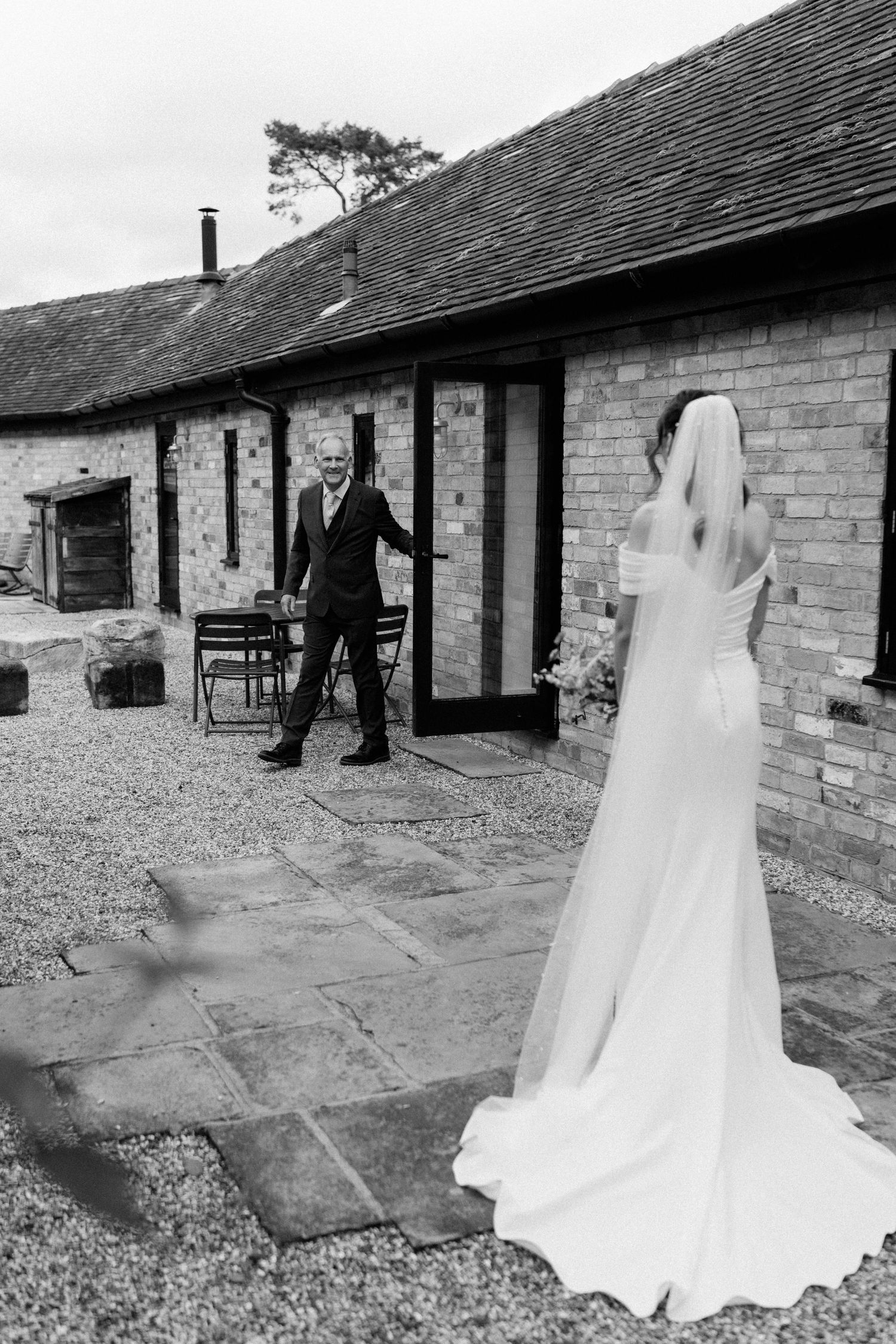 Groom reacting with joy as bride in white dress and veil approaches at rustic barn venue.