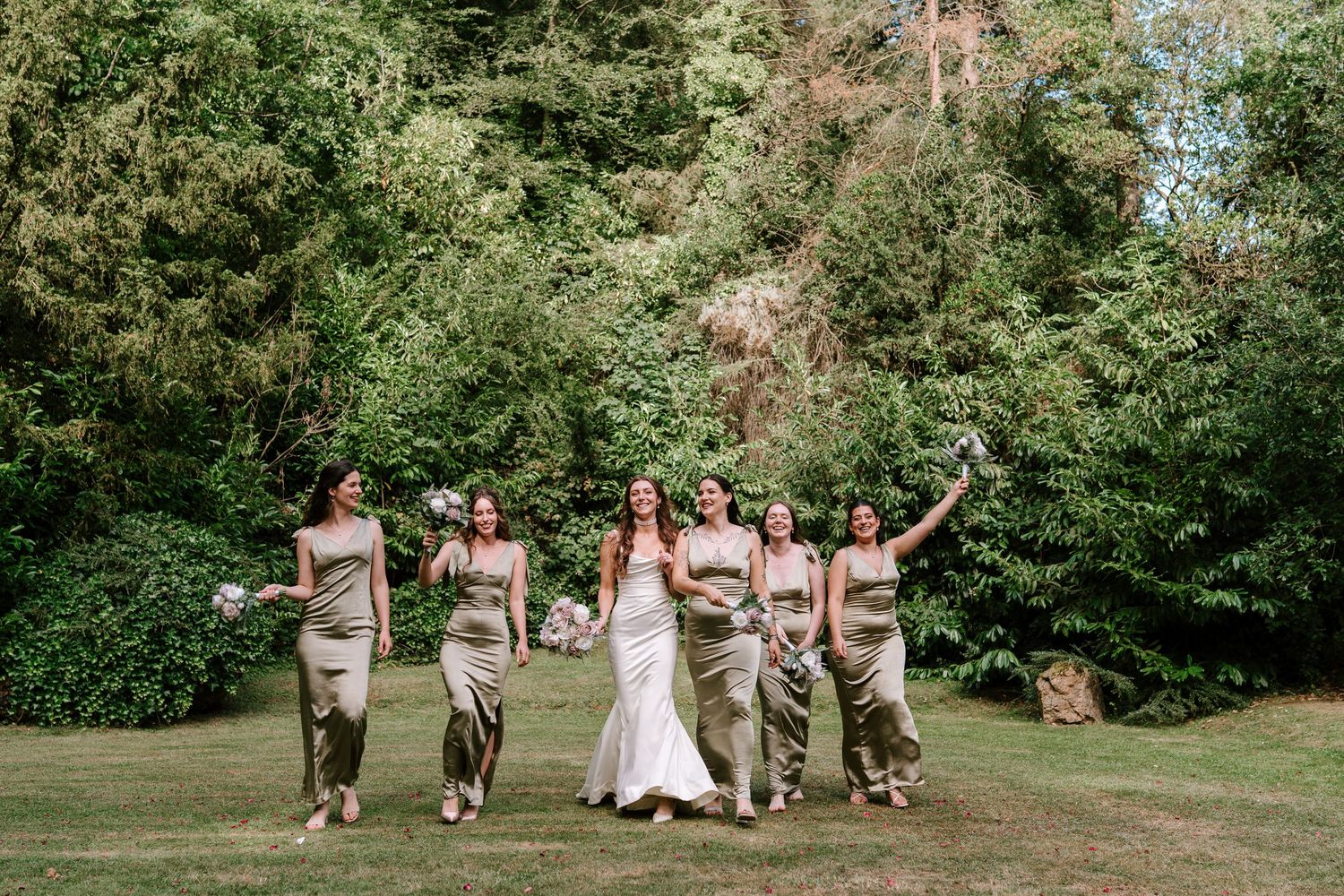 Bride in white gown walking joyfully with bridesmaids in gold satin dresses across a green lawn.