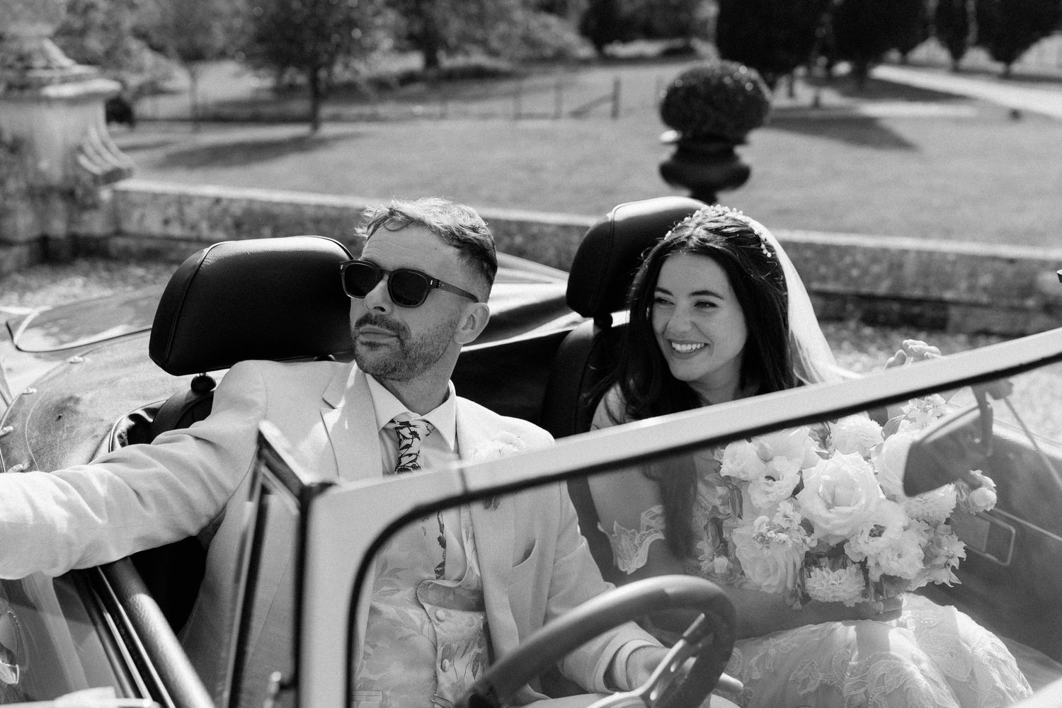 Bride and groom riding in a vintage convertible car on their wedding day, black and white photo.