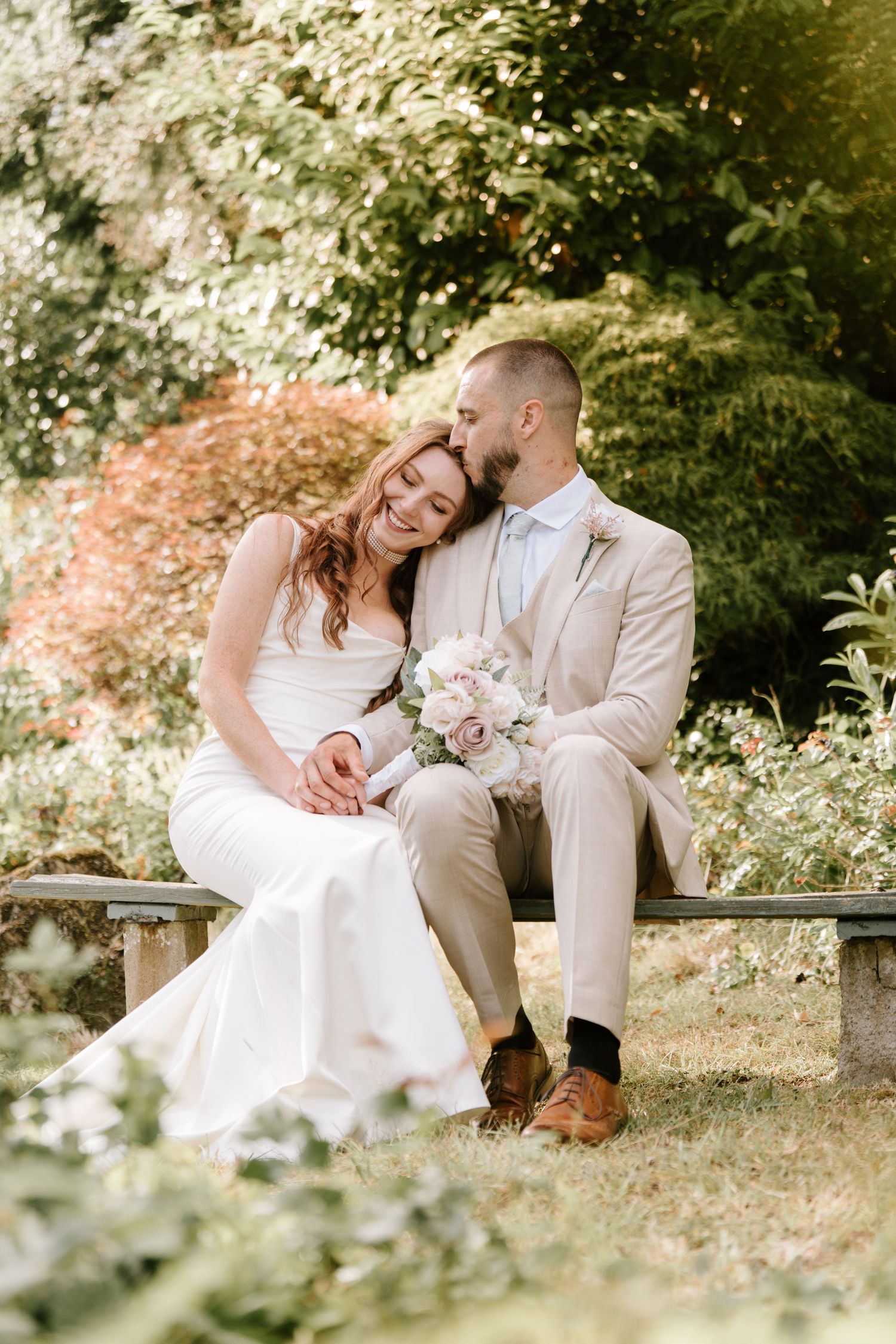 A smiling bride in white holds a bouquet while a groom in beige suit kisses her head on a garden bench.