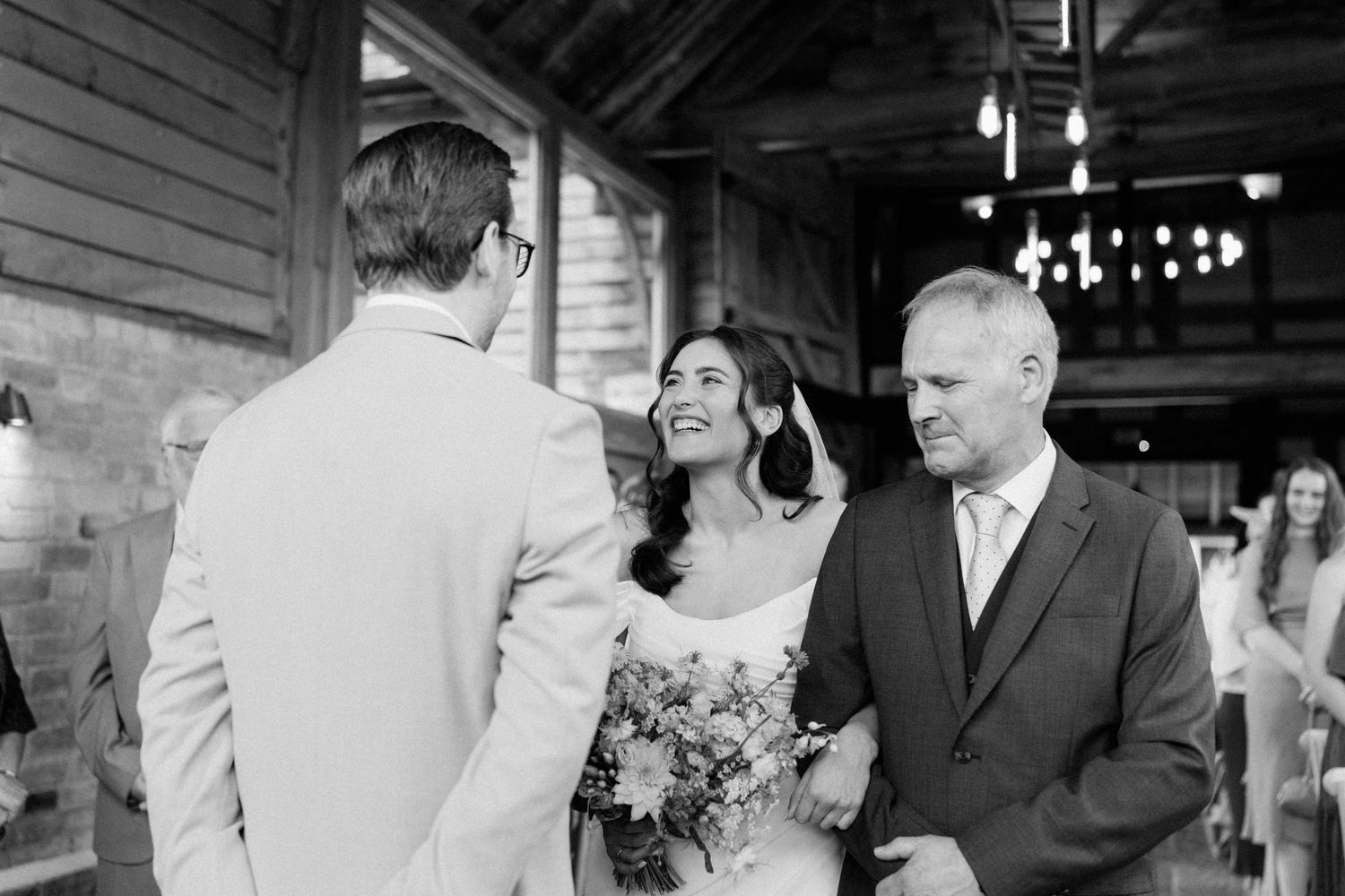 Black and white wedding photo of bride smiling at groom while father of bride looks on in rustic venue.