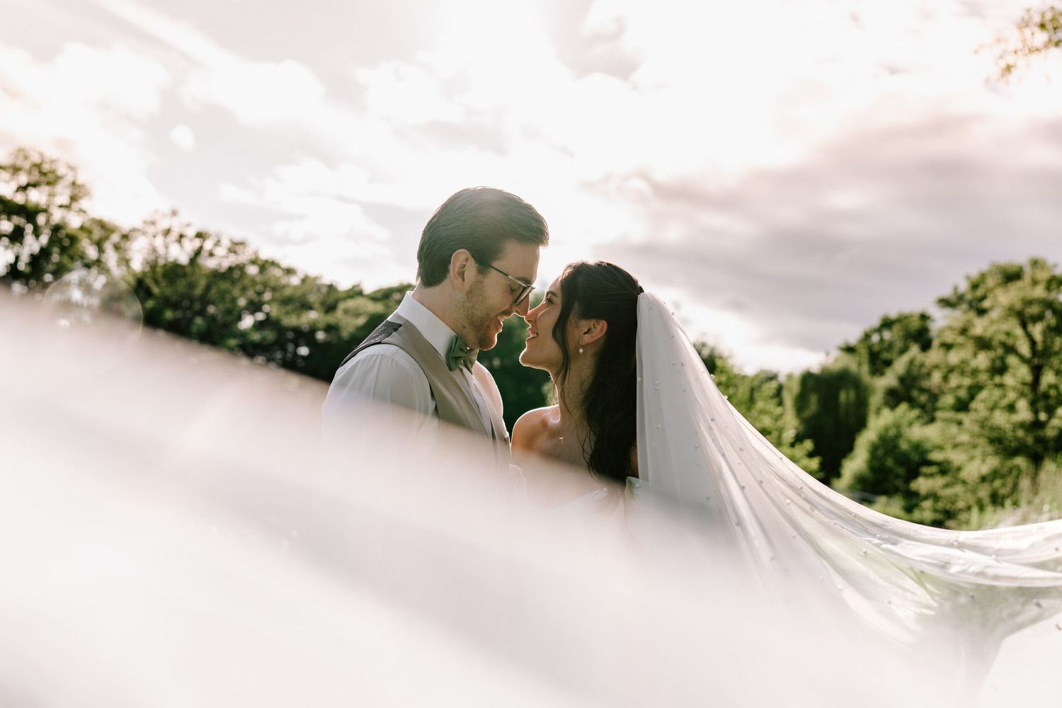 Romantic bride and groom touching foreheads with veil flowing in golden sunlight.