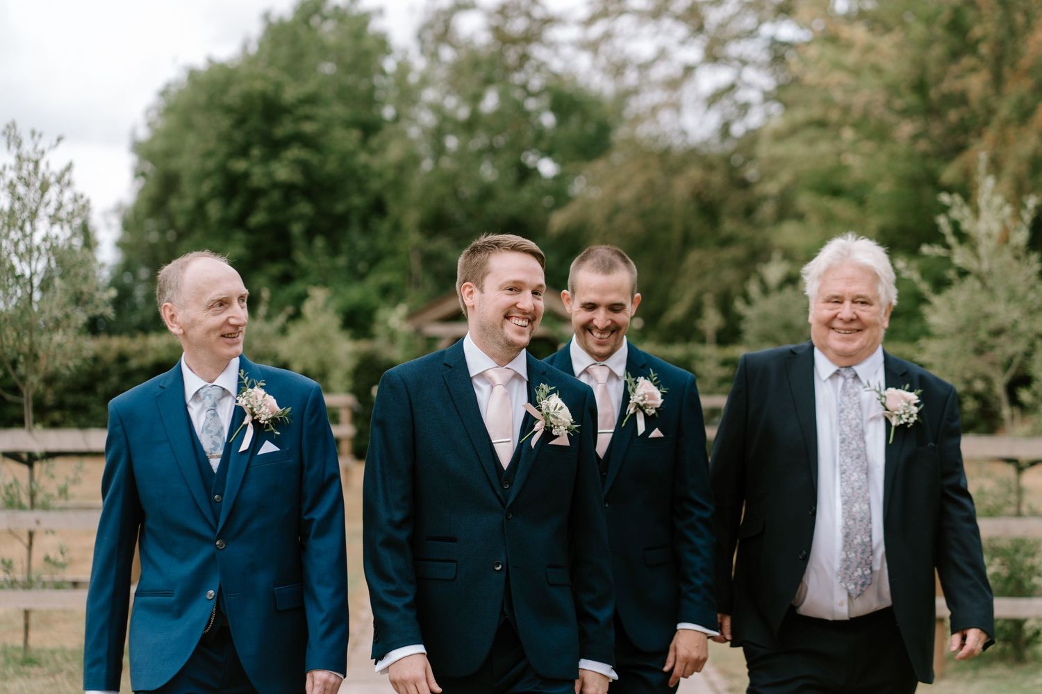 Four groomsmen in navy suits with floral boutonnieres walking together outdoors at a wedding celebration.
