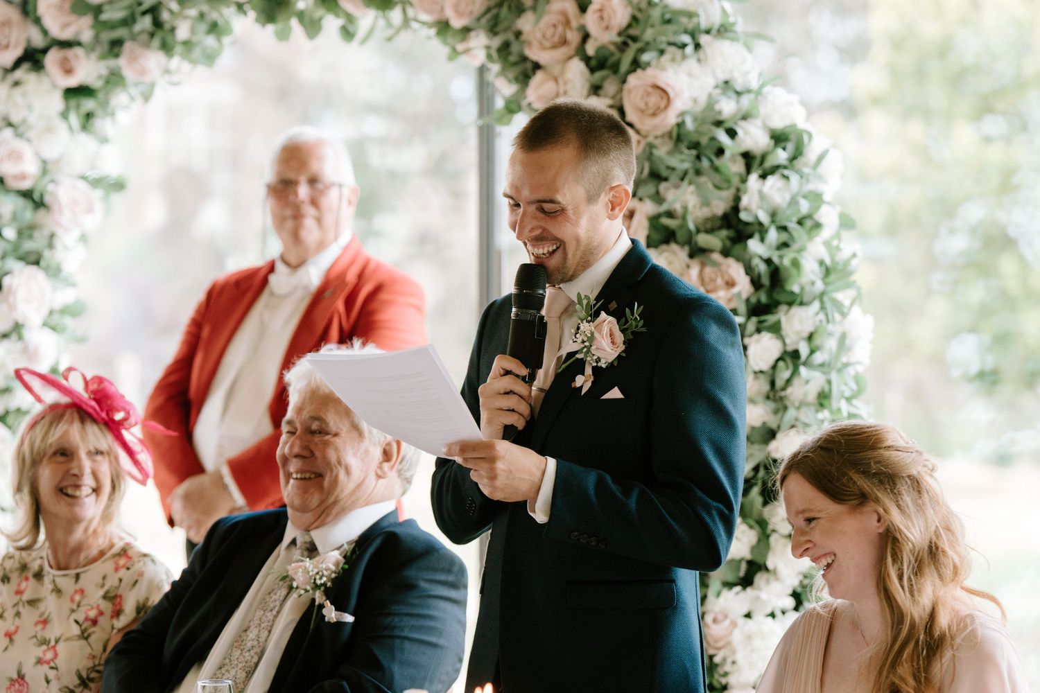 Groom giving a speech at wedding reception with floral backdrop, guests laughing and smiling in elegant attire.