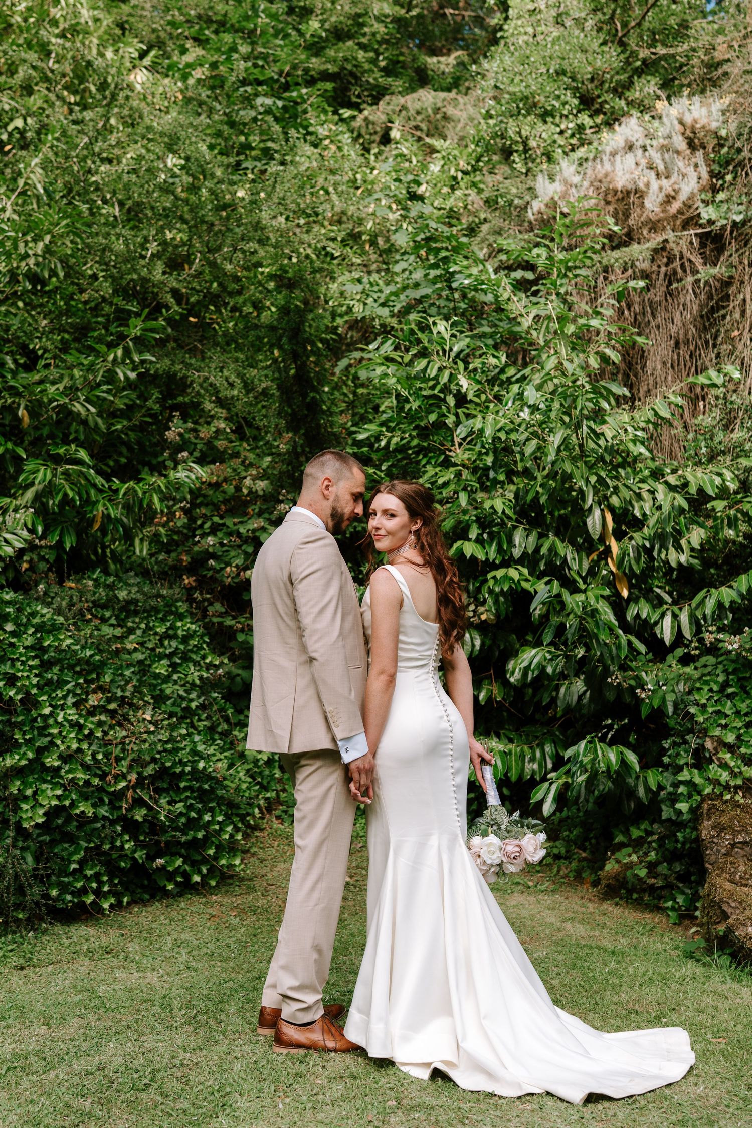 Bride and groom pose together in a lush green garden, bride holding a bouquet in elegant white gown.