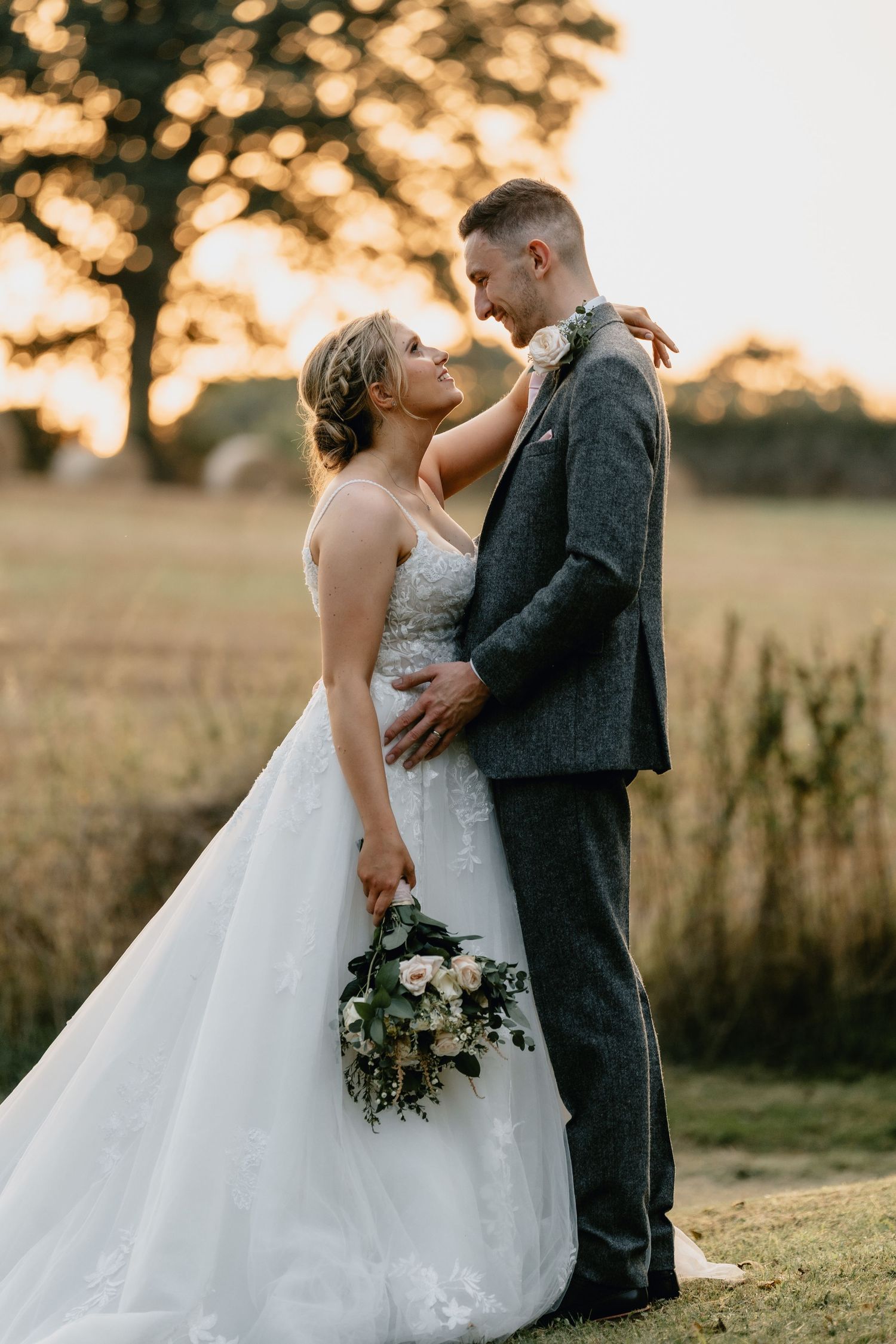 Bride and groom share a romantic golden hour moment in a field, bride holding a white floral bouquet in a lace gown.