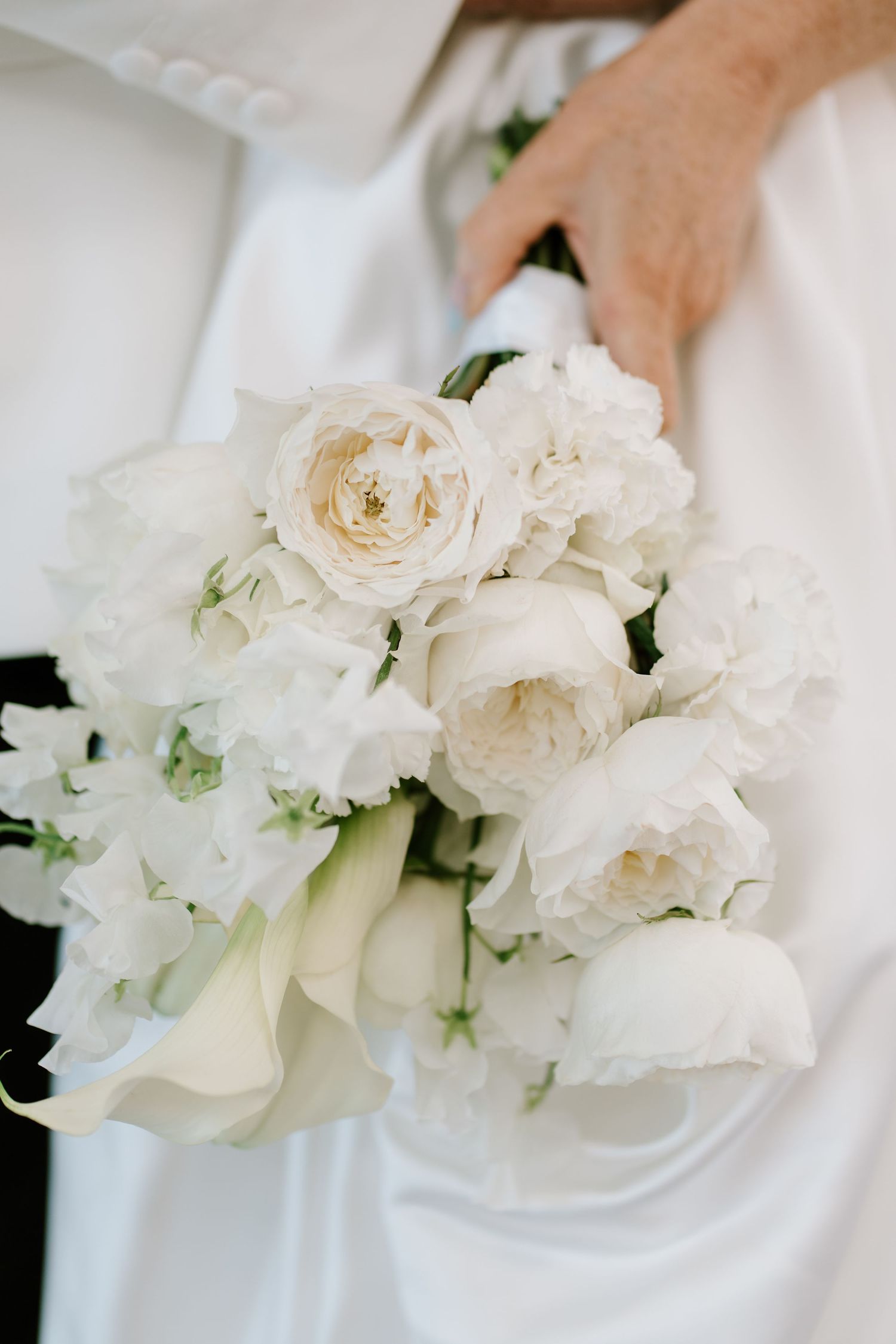 Bride holding a lush white wedding bouquet of roses, peonies, and calla lilies against a white satin gown.