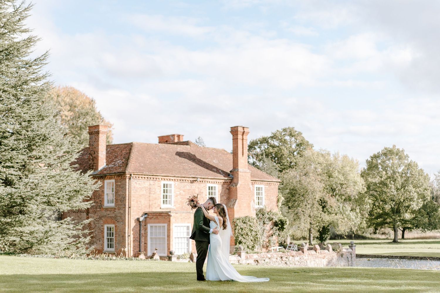 Bride and groom share a romantic moment on lush green lawn before elegant red brick English country house wedding venue.