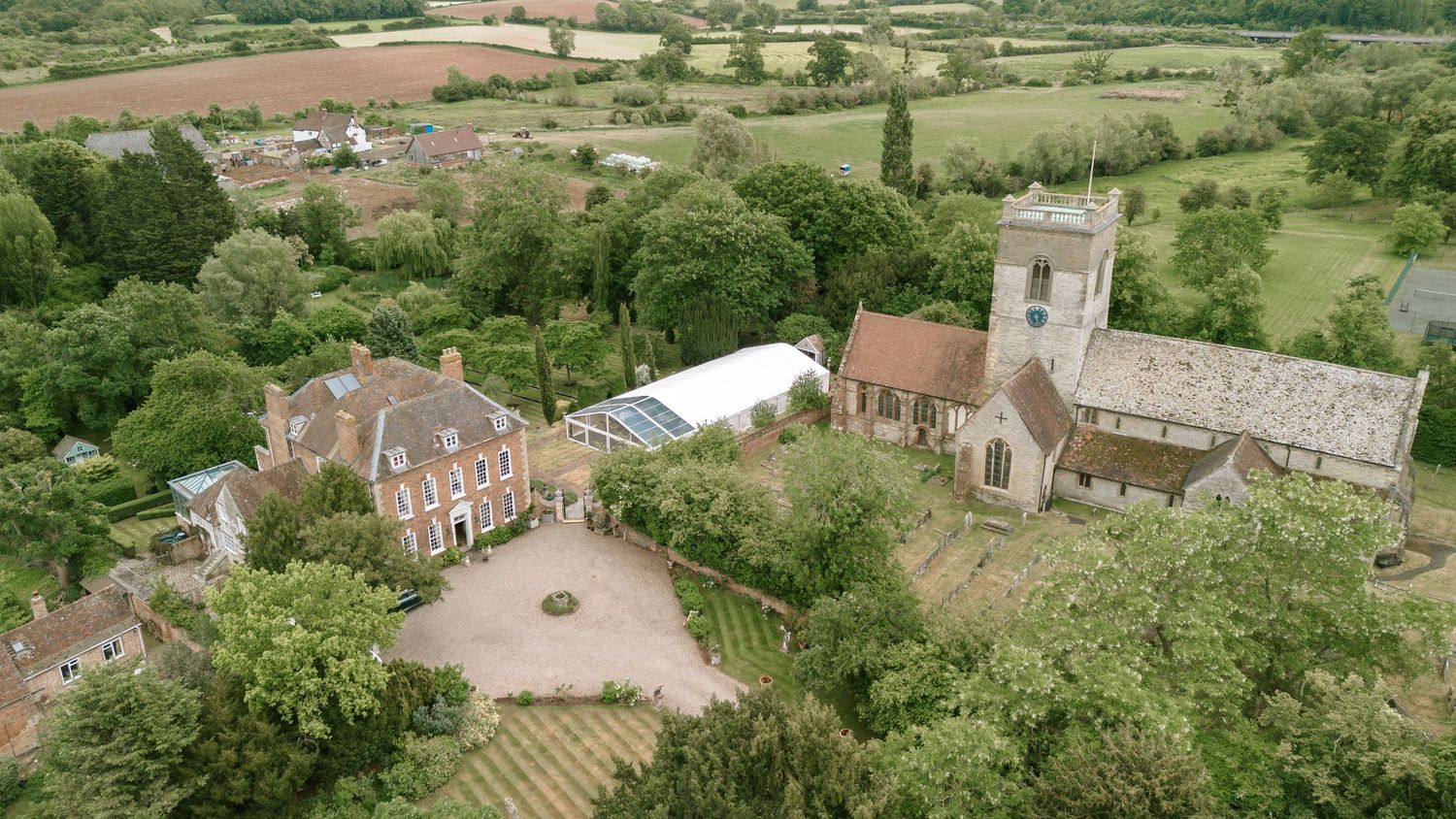 Aerial view of a historic English manor house and church surrounded by lush green countryside.