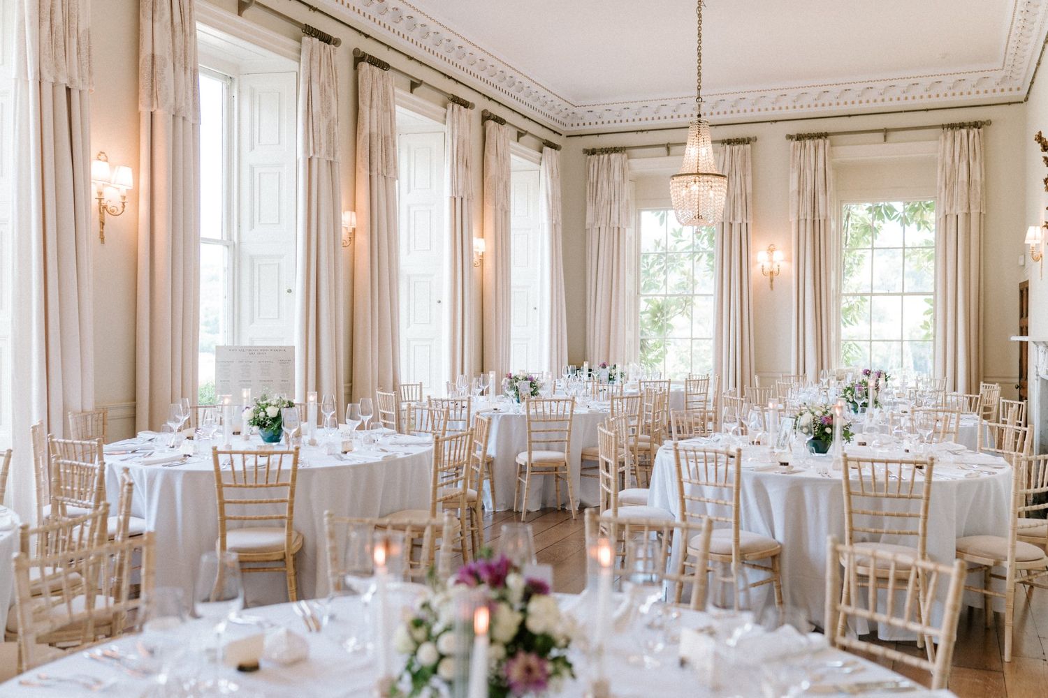 Elegant wedding reception hall with white round tables, gold Chiavari chairs, floral centerpieces, and tall draped windows.