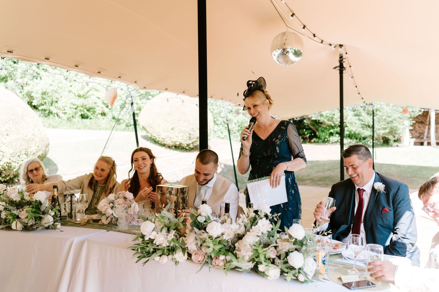 Wedding party seated at decorated head table under marquee during reception speeches.