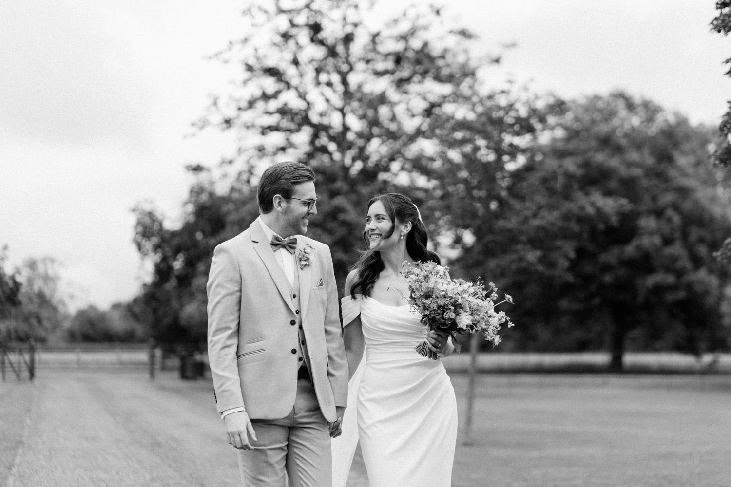 Black and white photo of bride and groom walking hand in hand on tree-lined path.