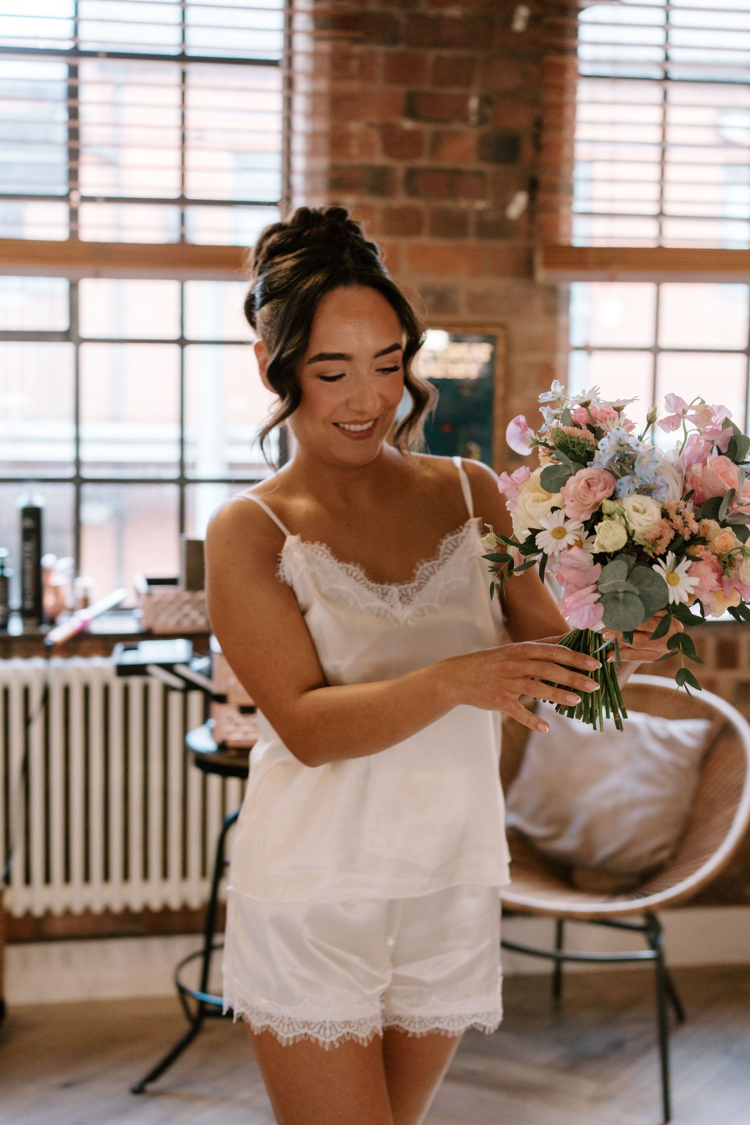 Smiling bride in white lace camisole set holding pink and white floral bouquet in rustic brick room.