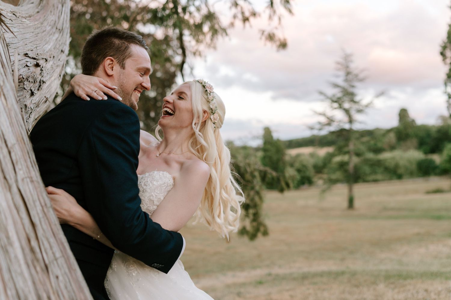 Happy bride and groom laughing together leaning against a tree in a lush green outdoor wedding setting.