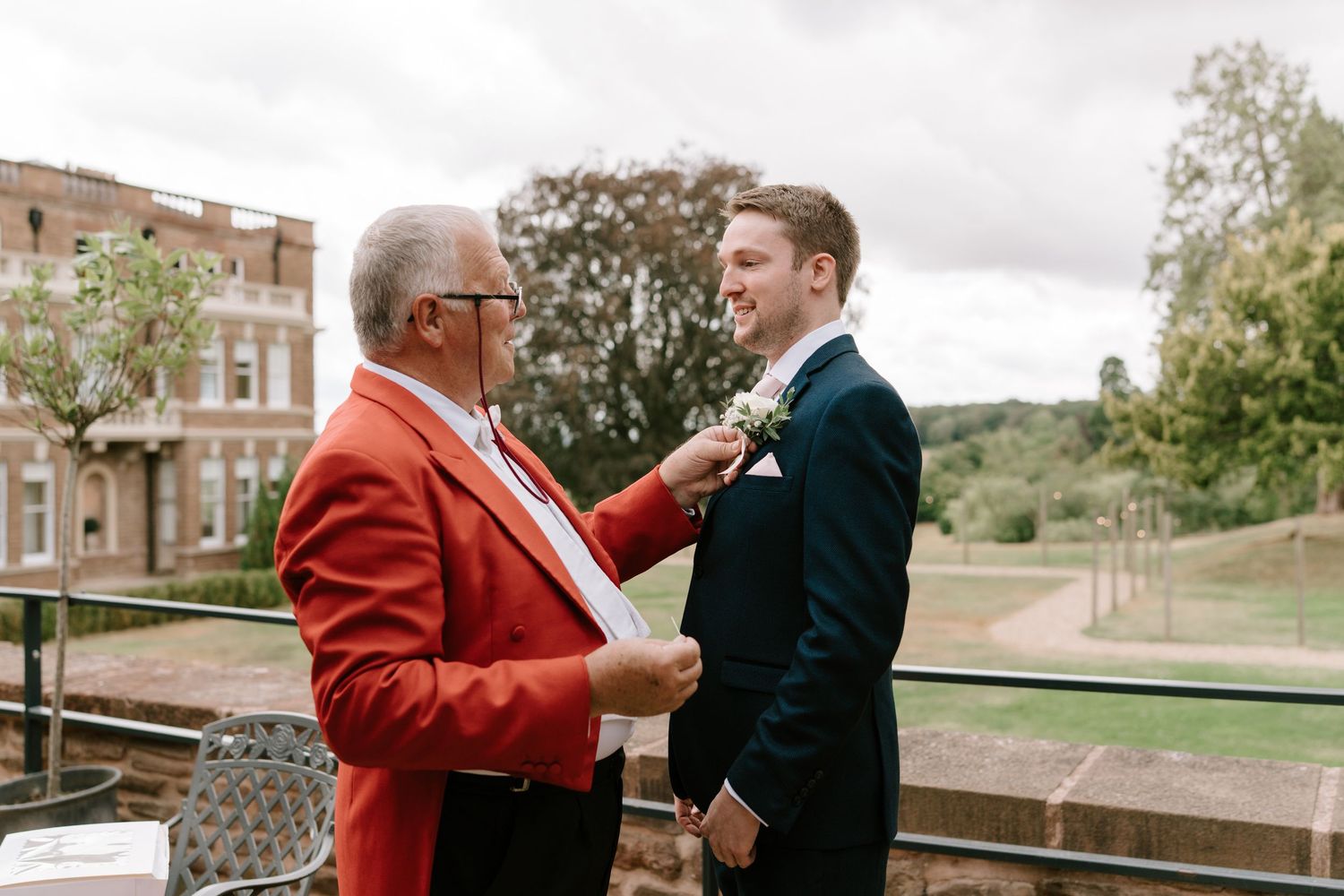 An older man in a red blazer pins a boutonniere on a groom in a navy suit at an outdoor wedding venue.