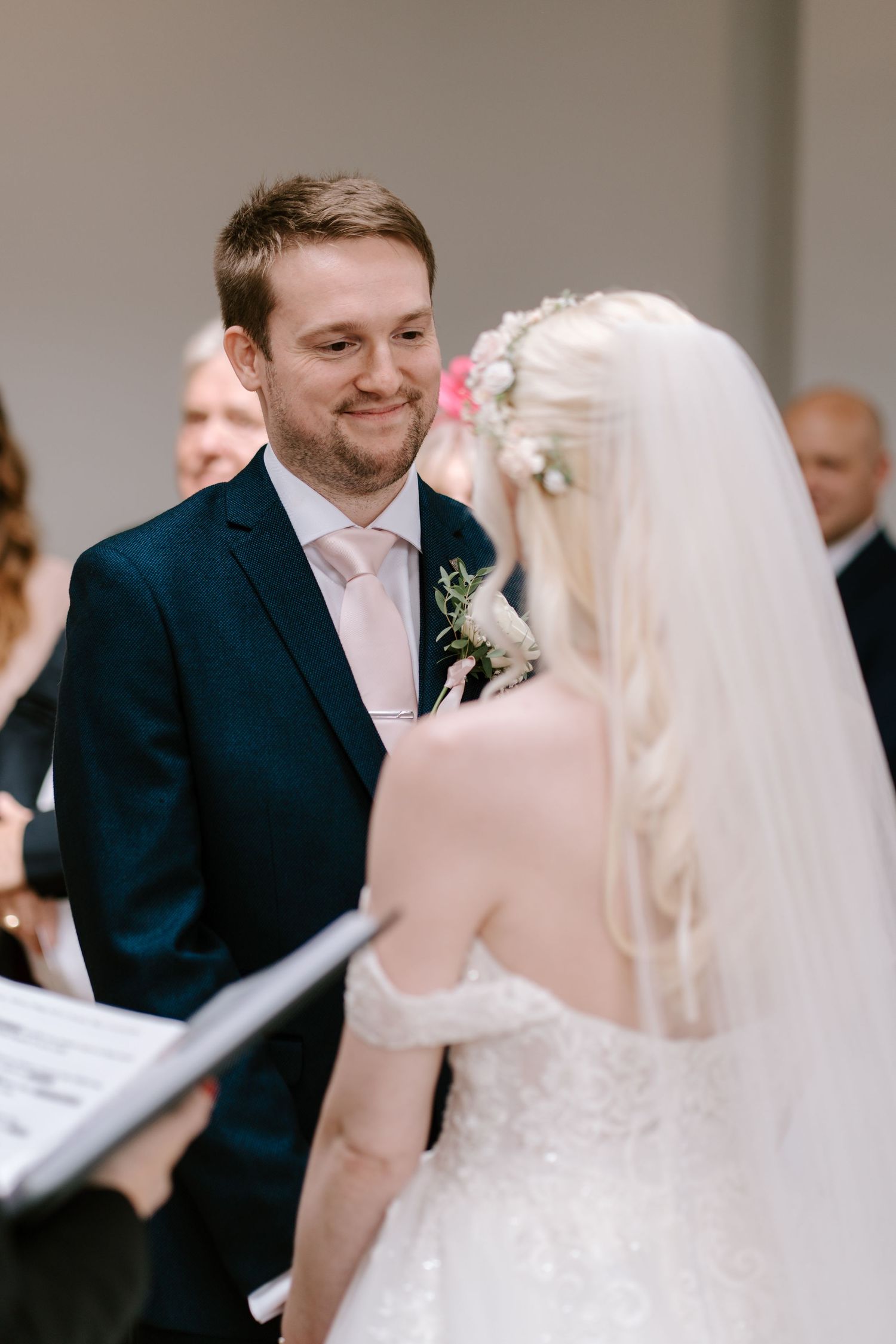 Groom smiling at bride during wedding ceremony, wearing navy suit with pink tie and boutonniere.