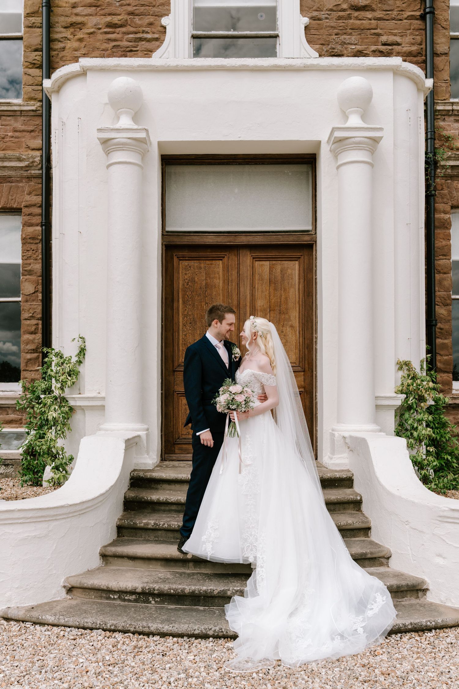 Bride and groom standing on stone steps before elegant white portico entrance of historic brick wedding venue.