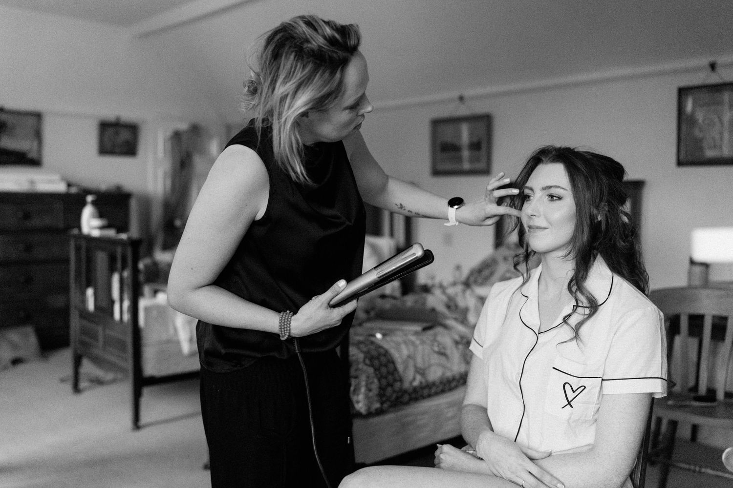 A makeup artist applies makeup to a bride-to-be during wedding preparations in a softly lit bedroom.